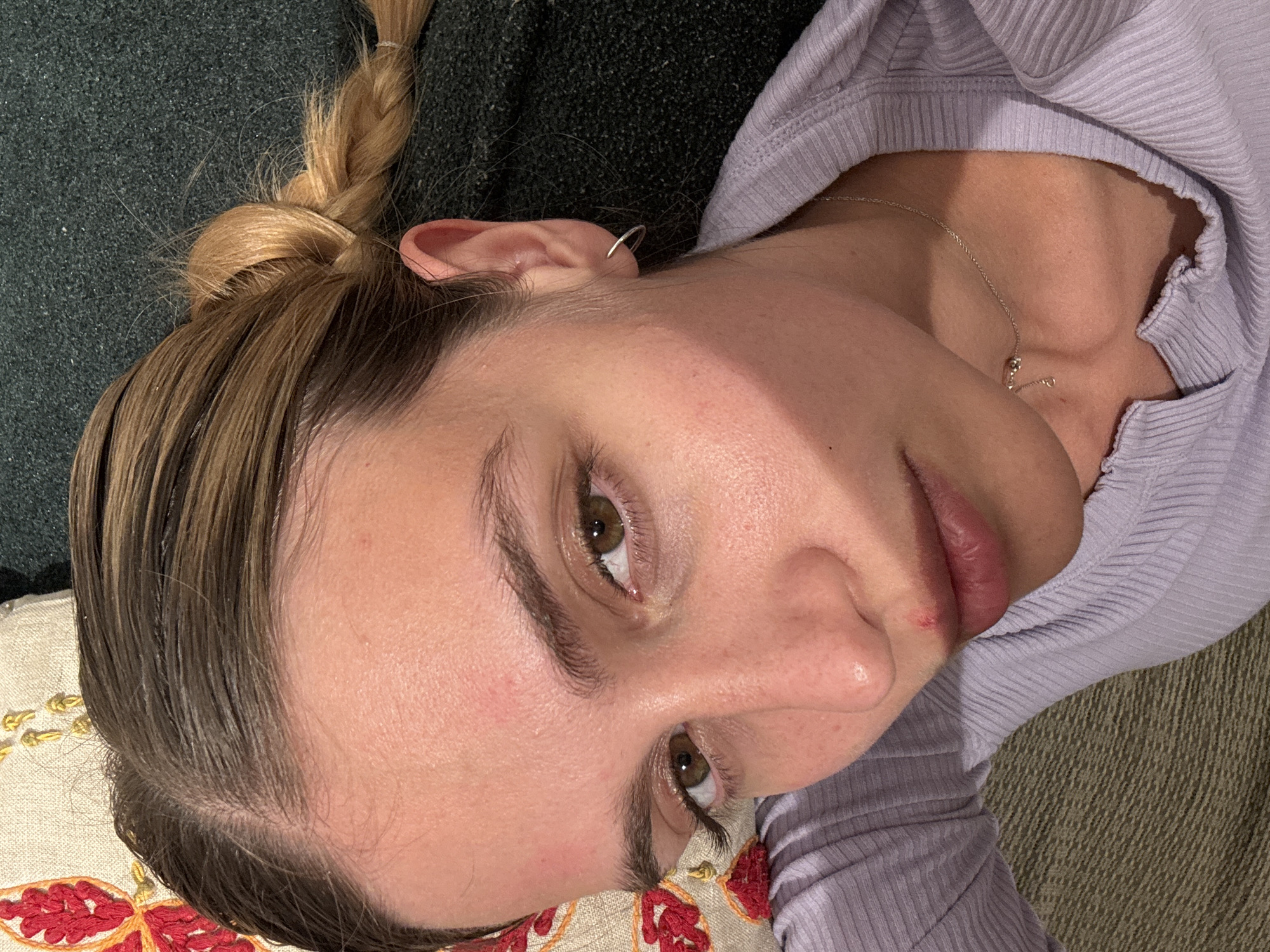 A close-up, high-angle shot captures a person lying down, looking directly at the camera. The subject, appearing to be female, is positioned with their head on the left side of the frame, tilted slightly. They have light brown hair with blonde highlights, pulled back into a visible braid on the top left. A small silver cuff earring adorns their right ear.
The person is wearing a light purple, ribbed long-sleeved top or hoodie, and a delicate silver chain necklace with a small pendant is visible around their neck and chest. Their complexion shows natural skin texture, and their brown eyes are open, meeting the viewer's gaze. A small reddish mark or sore is visible on the upper lip/philtrum area.
The background consists of a dark green, textured fabric (likely a couch or blanket) behind their head and a cream-colored cushion with red and gold embroidered patterns beneath their body. The lighting is soft, indicating an indoor setting. The overall orientation of the photo is rotated, presenting the subject from an overhead perspective while they are reclined. No visible text is present in the image.