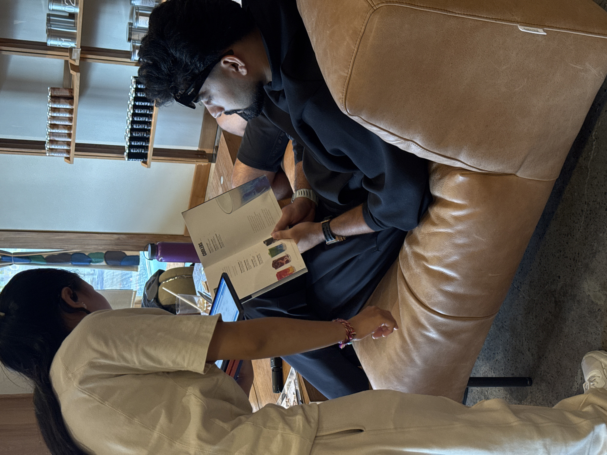 Inside a brightly lit café in North Kuta, Indonesia, two individuals are seated. A man in a black hoodie, sporting sunglasses on his head and a beard, sits on a tan leather couch, intently studying a menu. A woman wearing a white outfit stands beside him, looking at a laptop, possibly assisting him with a selection. A water bottle is visible near the window behind the woman. In the background, shelves display rows of canned goods.