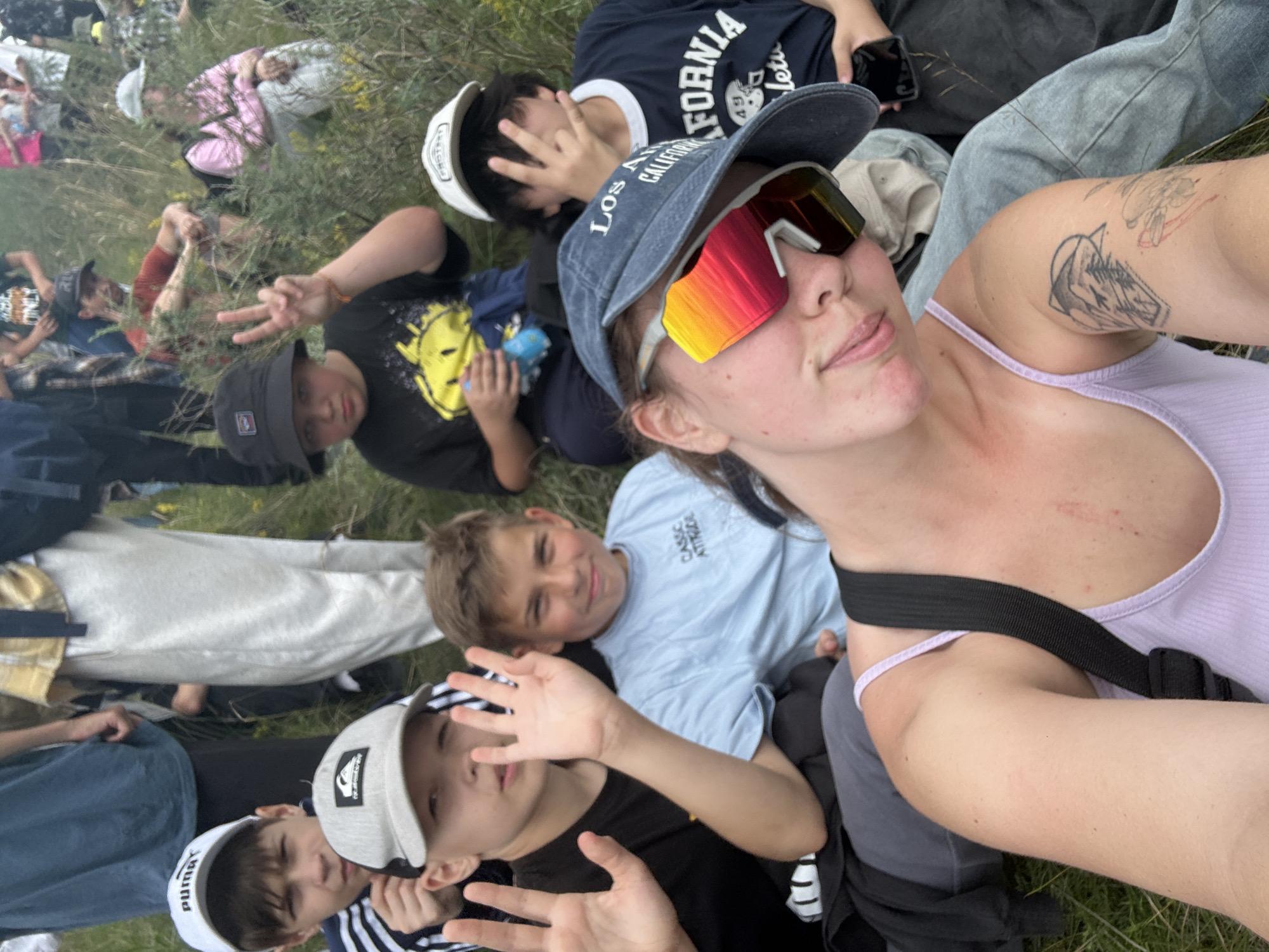 In this eye-level, outdoor, slightly tilted, selfie-style photograph, a group of individuals, appearing to be mostly young people, are gathered on grassy terrain. The subjects are positioned in a staggered, somewhat chaotic manner, with some lying down and others seated or standing. The scene suggests a casual outdoor gathering, possibly a picnic, festival, or simply a group of friends enjoying time together in a natural setting.
In the foreground, a young person wearing a blue cap with "LOS ANGELES CALIFORNIA" visible and reflective sunglasses with a vibrant, multi-colored lens is taking the selfie. They are wearing a light purple tank top. A black strap crosses their chest. Tattoos are visible on their left arm. To their left, a young person in a black t-shirt with a yellow smiley face graphic is looking towards the camera, with their hand raised in a peace sign. Next to them, a boy in a light blue t-shirt with "CLASSIC ATTITUDE" written on it is smiling and looking upwards. Another young person in a black and white striped shirt and a white cap with "PUMA" is also visible, with their hands in the air. Further in the background, other individuals are seen lying in the grass, with one person wearing a pink shirt and white pants.
The lighting suggests it is daytime, with a bright, slightly overcast sky. The overall mood appears to be relaxed and joyful, with smiles and gestures of peace and greeting. The presence of grass and foliage indicates a rural or park-like setting. While the specific event is not explicitly clear, the informal nature of the gathering and the relaxed poses suggest a leisurely outdoor activity. There are no specific visual cues to suggest the time of day or the weather conditions beyond it being daylight and outdoors. The location context provided mentions Nikolo-Petrovka, Russia.