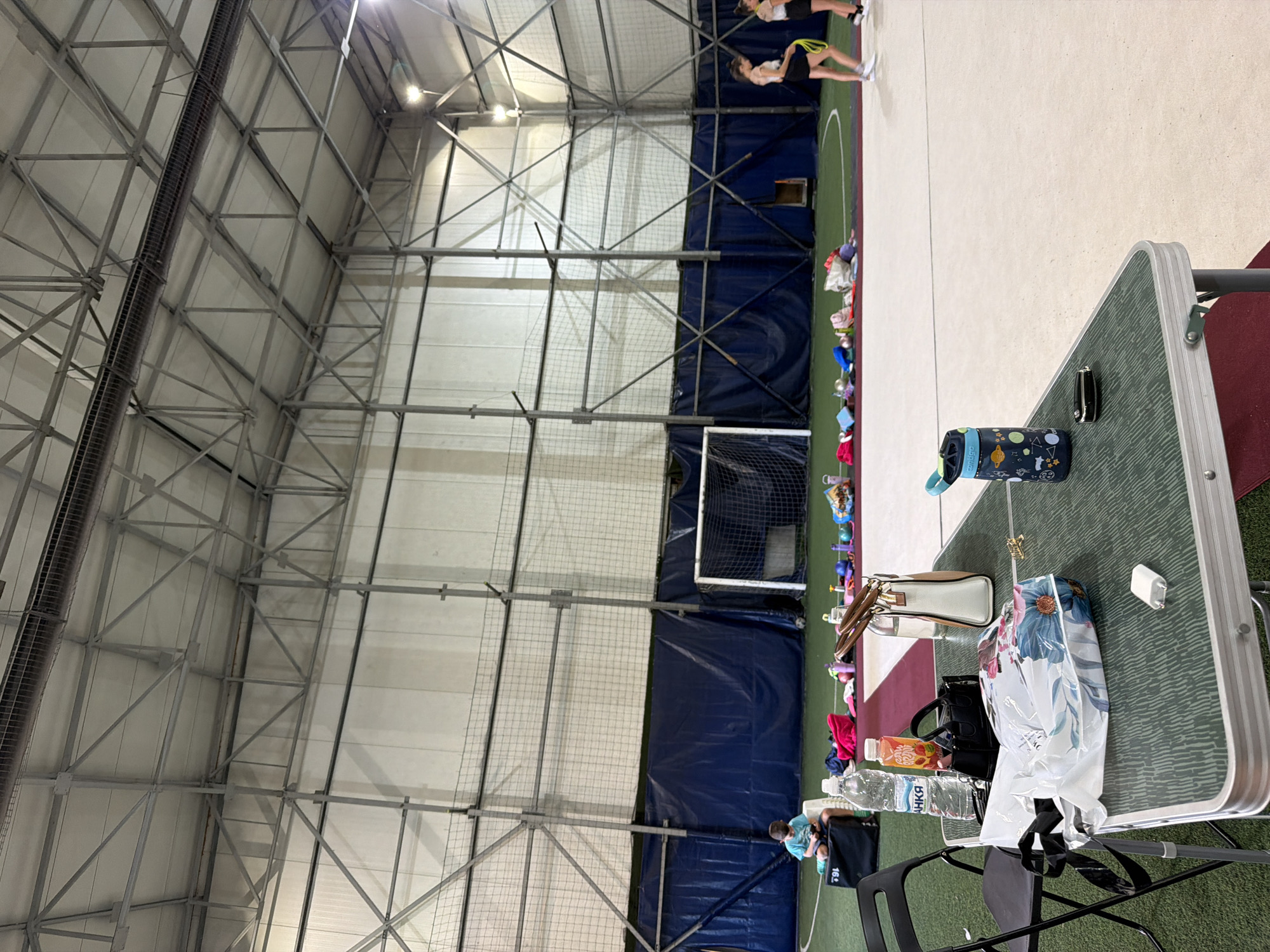This image depicts the interior of a gymnasium, likely at the National Gymnastics Training Center in Sofia, Bulgaria, given the presence of rhythmic gymnastics equipment. The scene is set on a green artificial turf floor, with a white border demarcating a training area. The ceiling features a complex metal truss system, and large blue tarpaulins cover the walls.
In the foreground, a green and silver folding table is set up, laden with various items. These include a large white plastic bag with a floral print, two small black handbags, a white and tan handbag, a blue water bottle with a patterned design, a bottle of water, and a bottle of juice with a colorful label. A pair of white earbuds is also on the table. To the left of the table, a black folding chair is positioned.
In the middle ground, several individuals are visible. A young boy in a blue t-shirt sits on a black bag on the left side of the frame. Further to the right, a collection of rhythmic gymnastics apparatus, including ribbons, balls, and bags, are laid out on the floor. Two young women, dressed in athletic attire, are standing in the background on the right, one of whom is holding a yellow ribbon. A white soccer goal with a net is positioned against the far wall.
The lighting suggests an indoor environment, possibly late afternoon or evening. The overall atmosphere is one of readiness for or a break during a sports training session.