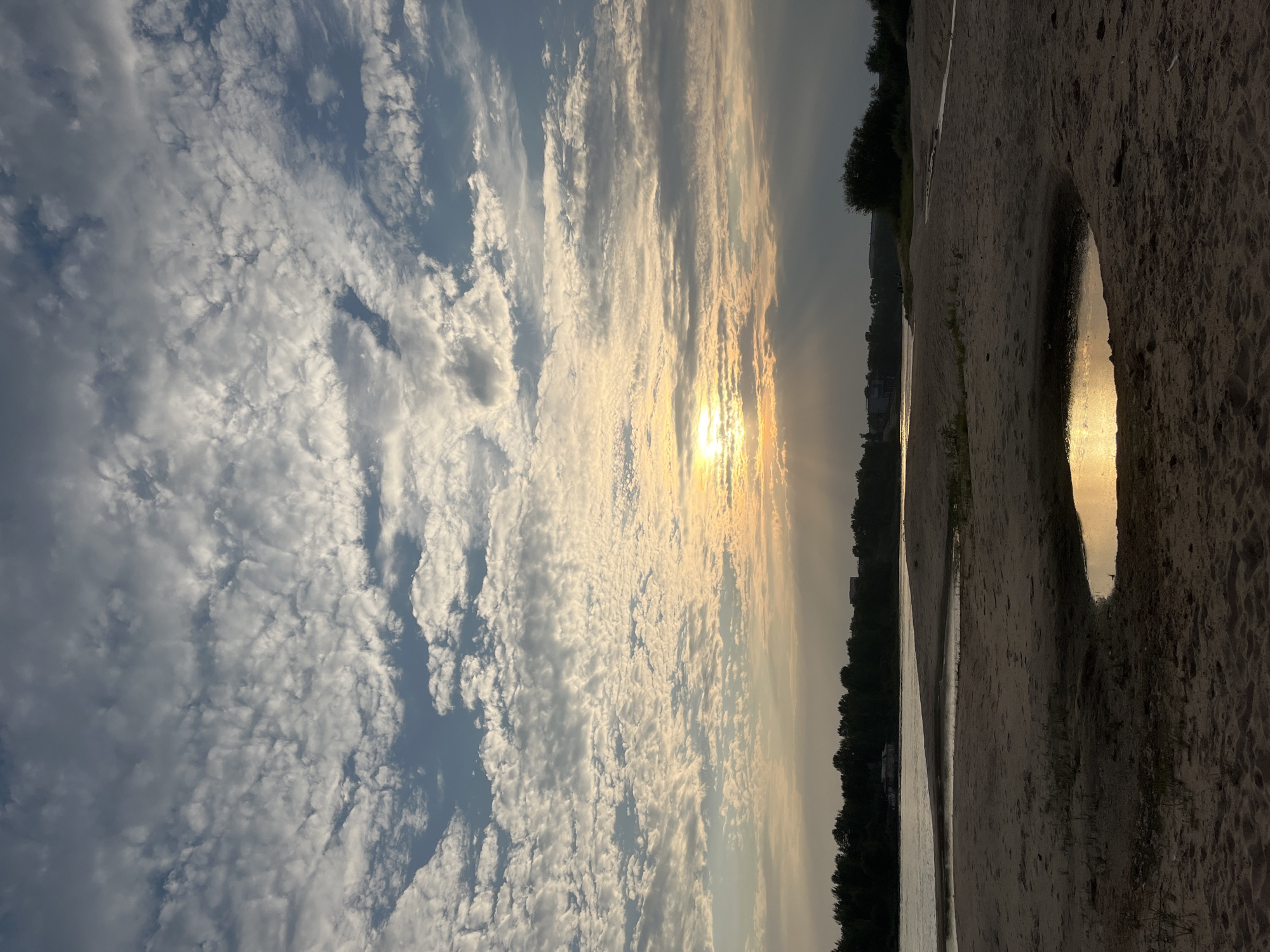 The image depicts a sunset over a sandy beach and a river, likely the Volga River, in Nizhny Novgorod, Russia. The sky is filled with scattered, wispy clouds, with the sun partially obscured by them, casting a warm, golden light and visible sunbeams. In the foreground, a sandy beach stretches out, with a small pool of water reflecting the sunset. A gentle slope leads down to the river, which appears calm. Across the river, a dense line of trees forms the shoreline, with some buildings visible in the distance. The overall atmosphere is serene and natural. There are no people or animals visible in the image.