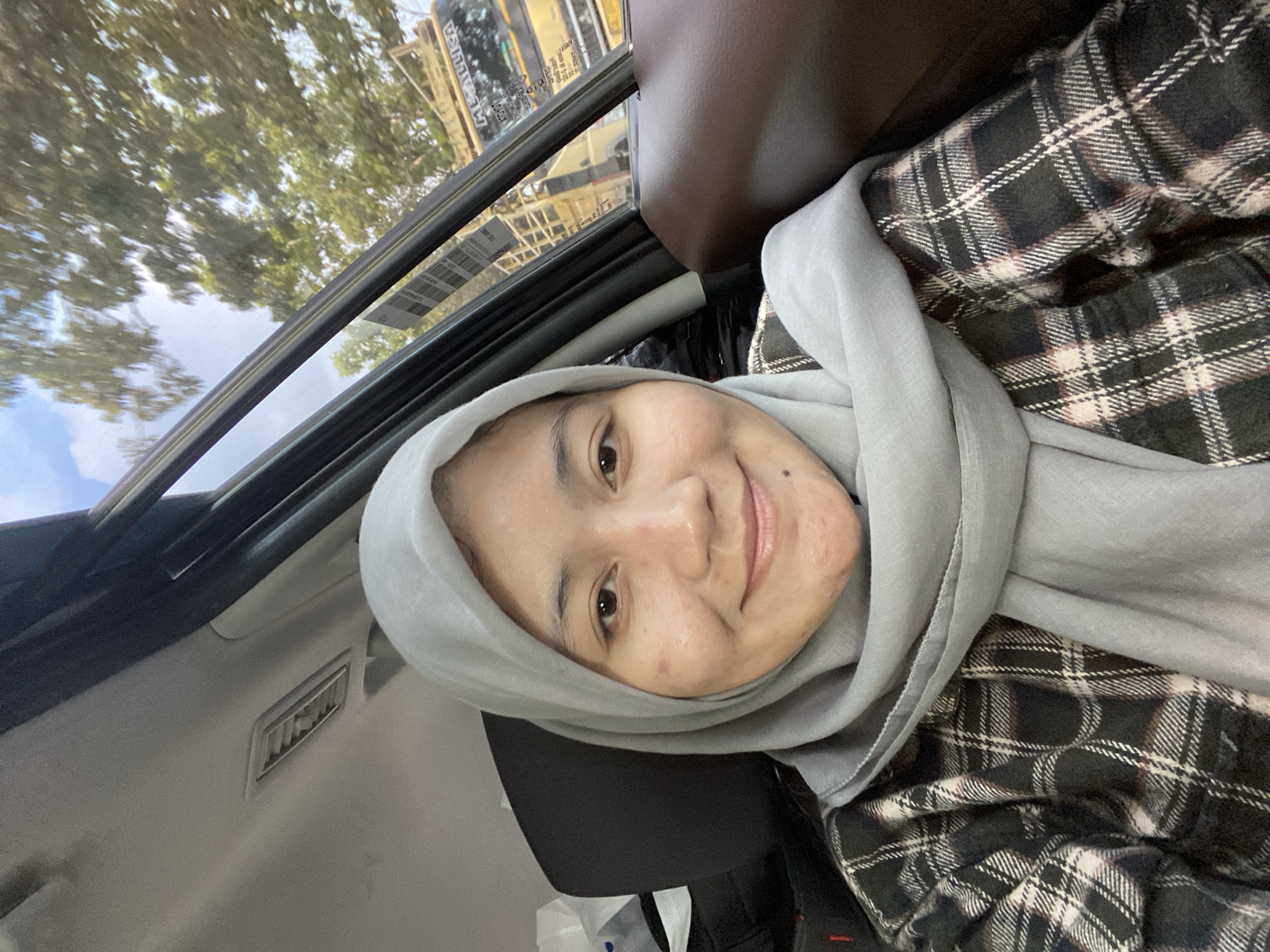 The image is a selfie taken inside a vehicle, likely in Gambut Barat, Indonesia, given the context. A young woman wearing a light grey hijab and a plaid shirt is the primary subject. She is smiling at the camera. The interior of the car is visible, including the headliner, air vent, and car door. Through the windows, trees and a yellow truck with the word "METALLIKA" visible are seen, suggesting an outdoor setting with traffic. The lighting indicates it is daytime.