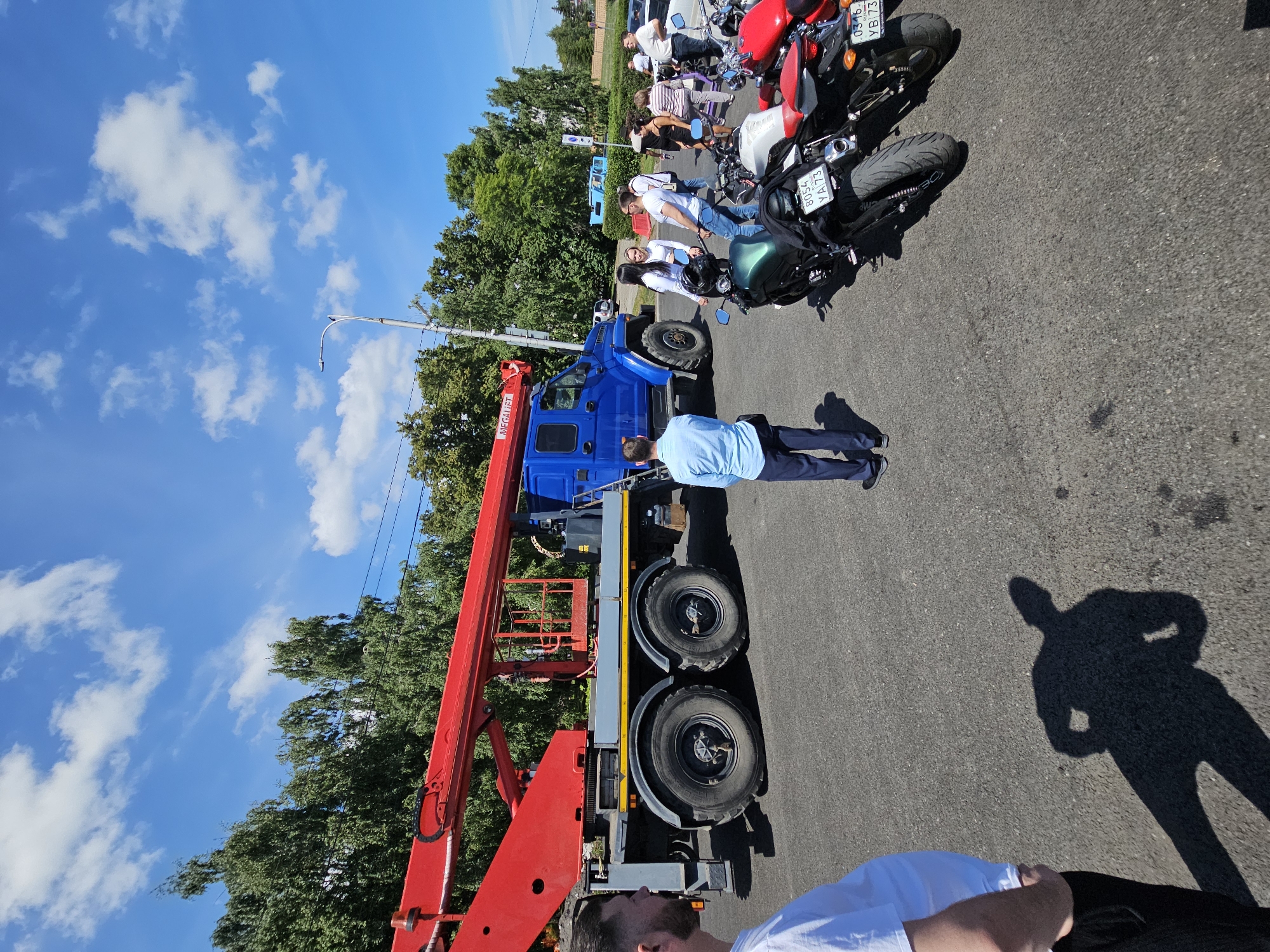 A bright, sunny day is depicted in this image, set against a backdrop of a clear blue sky with scattered white clouds. Trees line the background, suggesting an outdoor urban or suburban setting. In the foreground, a large blue truck with a red crane is the central subject. The crane is extended, with a cage-like platform at its end.
Several people are present around the truck and on the pavement. To the left, a man in a white t-shirt and dark pants faces away from the camera. In the center, a man in a light blue shirt and dark pants stands with his back to the viewer, facing the truck. A group of people, including women in white tops, are gathered near the rear of the truck, some interacting with each other.
Several motorcycles are parked on the asphalt in the mid-ground, with their license plates visible. The environment appears to be a paved area, likely a street or parking lot, with the shadow of a person cast on the ground in the lower left portion of the image. There is a street light visible in the background. The license plates on the motorcycles appear to be Russian. Based on the plate format and the visible Cyrillic script, the location is likely Ulyanovsk, Russia.