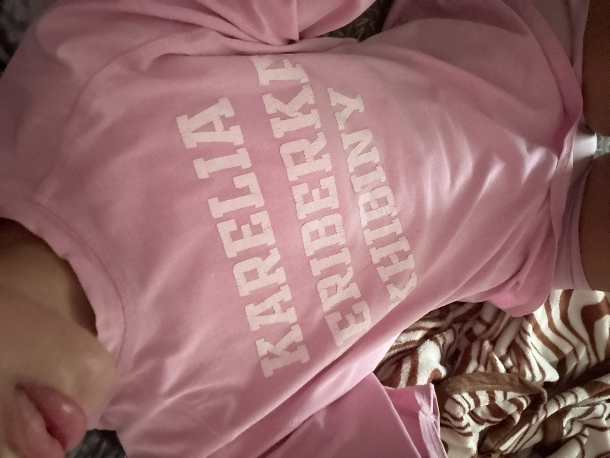 The image shows a close-up of a person wearing a pink t-shirt with white lettering that reads "KARELIA CHIBINY". The person's lips and part of their chin are visible at the top right of the frame, suggesting they are looking towards the camera. The t-shirt is draped over the person's torso and arm, with part of a blanket or patterned fabric visible in the background. There is no discernible indication of the city of Basarabeasca, Moldova, or any specific location within it.