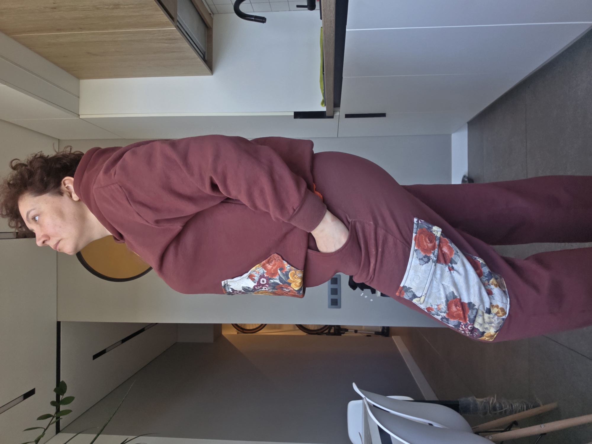 A woman with curly brown hair stands indoors in a burgundy hooded sweatshirt and matching sweatpants with floral patchwork details. She faces left with her hand in her pocket. The background reveals a modern interior with white cabinets, a wooden countertop, and a glimpse of a bicycle. A plant is visible in the lower-left corner. The scene suggests a contemporary living space. No landmarks or identifying features link it to Saint Petersburg.