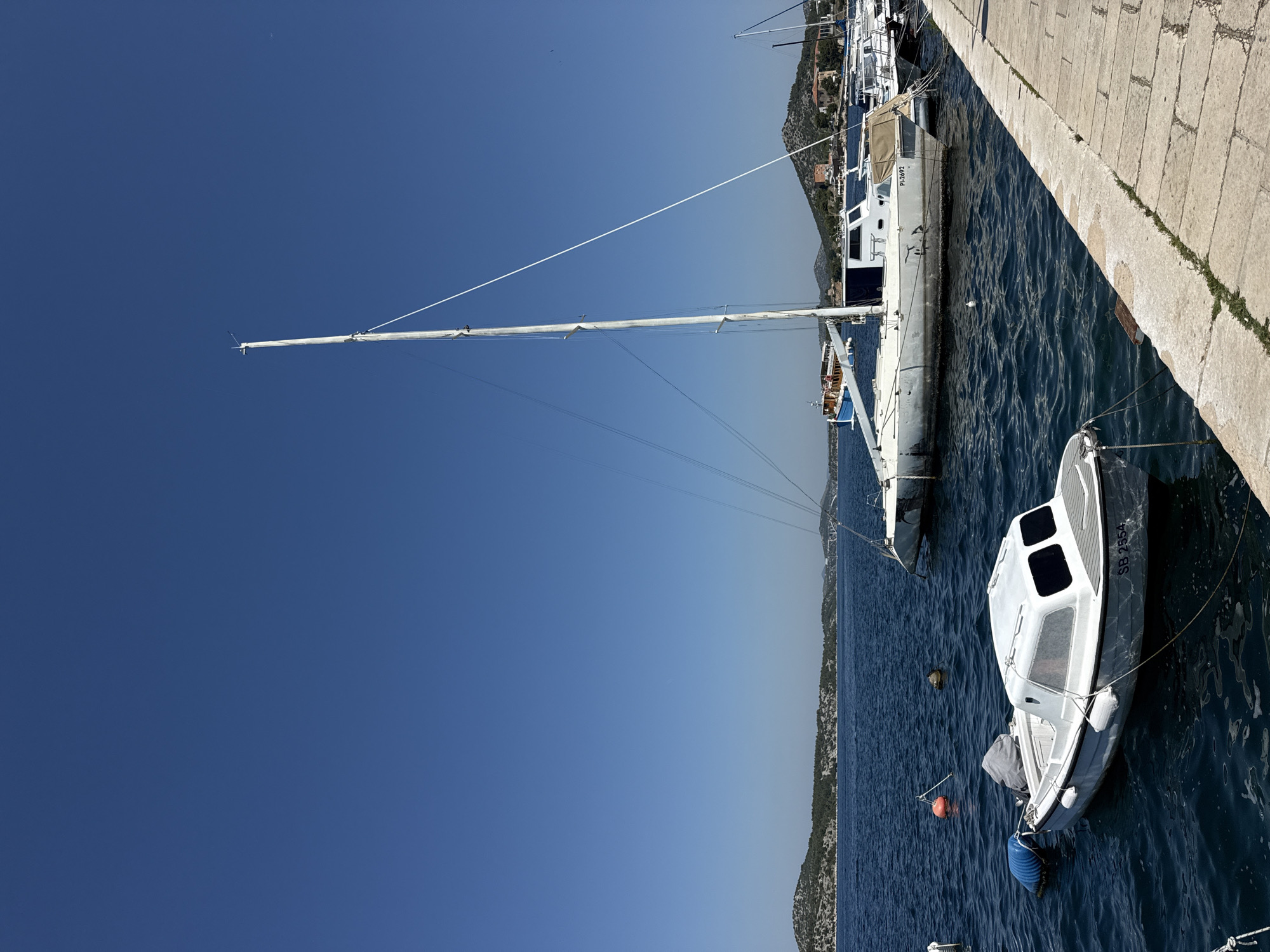 The image shows a bright, sunny day at a marina in Šibenik, Croatia. The water is a deep blue, and the sky is a clear, vibrant blue. In the foreground, a small white motorboat with a covered cabin is moored. The boat has the registration number "SB 2554" visible on its stern. To the right, a larger, older white sailboat with a tall mast is docked. Another sailboat, partially obscured, is visible behind it, along with other vessels. The marina has a stone-paved walkway along the right edge of the frame. In the background, a hilly, green coastline with scattered buildings and trees stretches across the horizon. The overall scene depicts a peaceful, picturesque coastal setting.