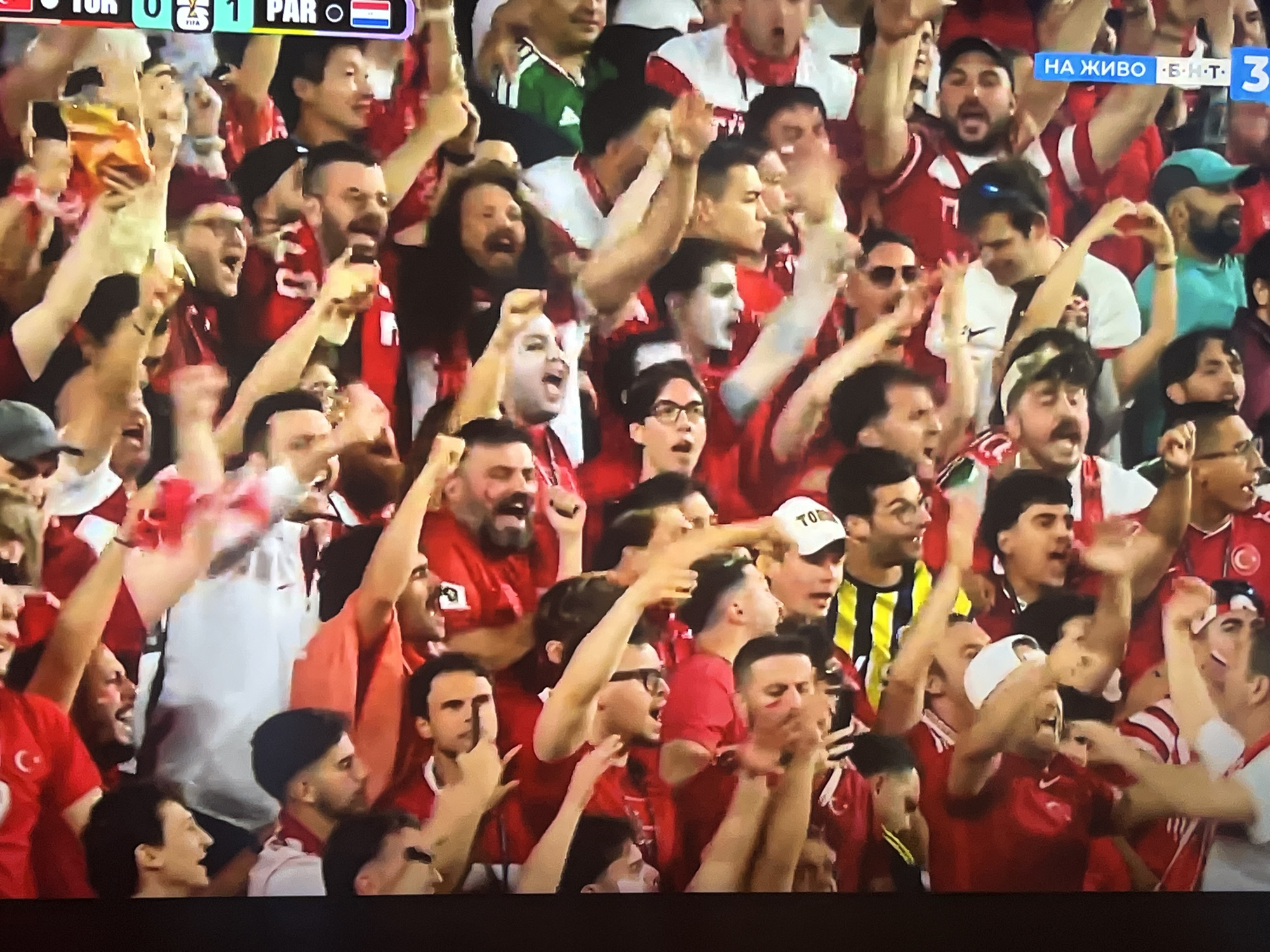 The image depicts a crowd of people, likely fans, in a stadium or viewing area. They are predominantly wearing red attire, some with white accents, and many are actively cheering or celebrating, with raised arms and open mouths. The atmosphere appears energetic and engaged, suggesting a sporting event or similar public gathering. The scoreboard at the top indicates "TUR 0 1 PAR", implying a football (soccer) match between Turkey and Paraguay. A blue banner with Cyrillic text "НА ЖИВО - БНТ 3" is visible in the upper right, indicating a live broadcast on Bulgarian National Television channel 3. Given this context, the location is likely a public viewing area in Varna, Bulgaria, where fans have gathered to watch the match.