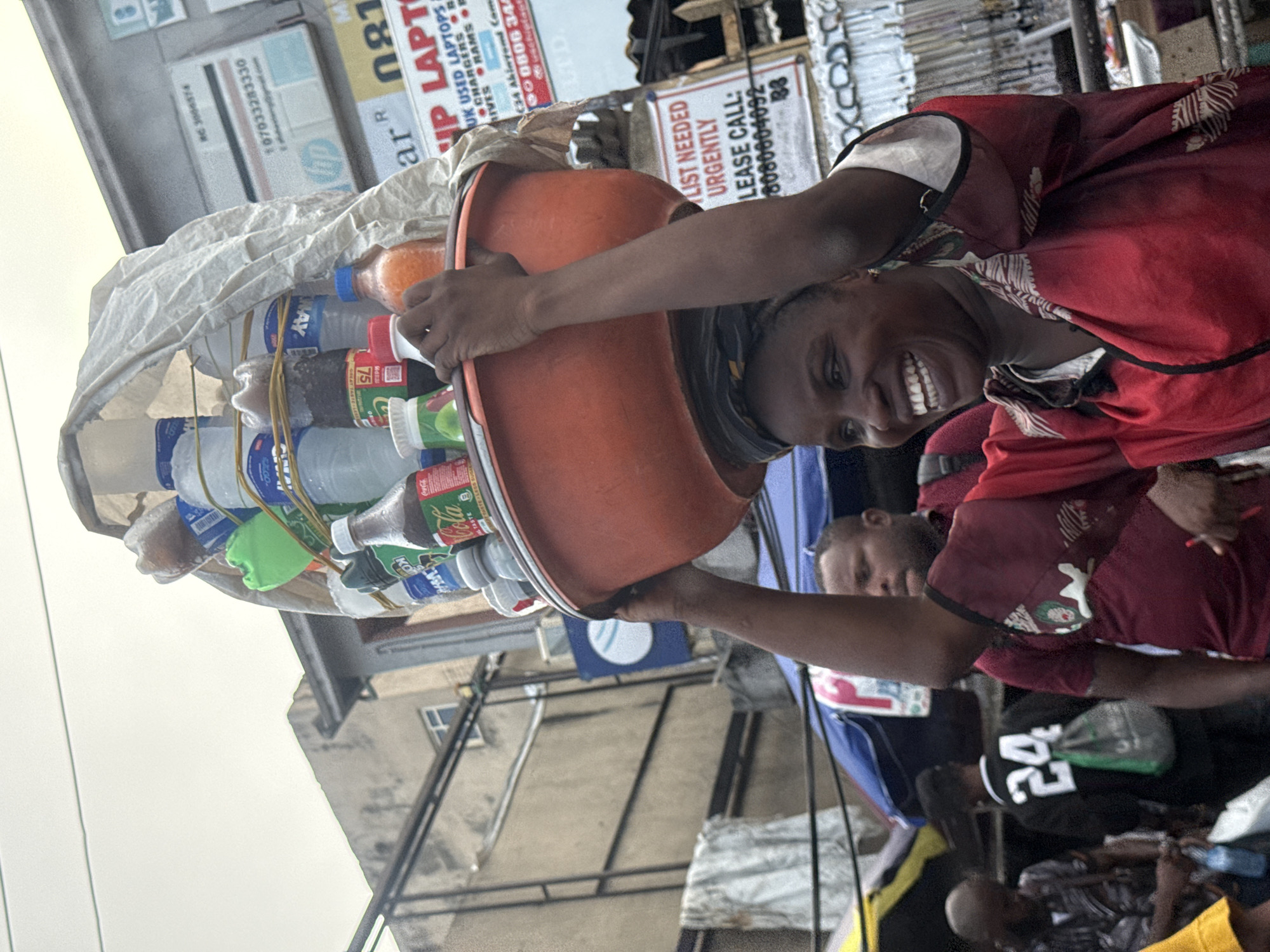 A woman with dark skin and braided hair, wearing a red and white patterned shirt, balances a large orange bowl filled with drinks and other items on her head. She is smiling widely, showing her teeth. The scene appears to be a busy street market, likely in Lekki, Nigeria, indicated by the signage visible in the background which includes advertisements for "HP USED LAPTOPS" and phone numbers. The background also shows buildings, awnings, and other people. The lighting suggests it is daytime, with an overcast sky. The woman is the central subject, and the focus is on her carrying the goods, which are mostly bottled beverages like Coca-Cola and other soft drinks, along with some plastic cups.