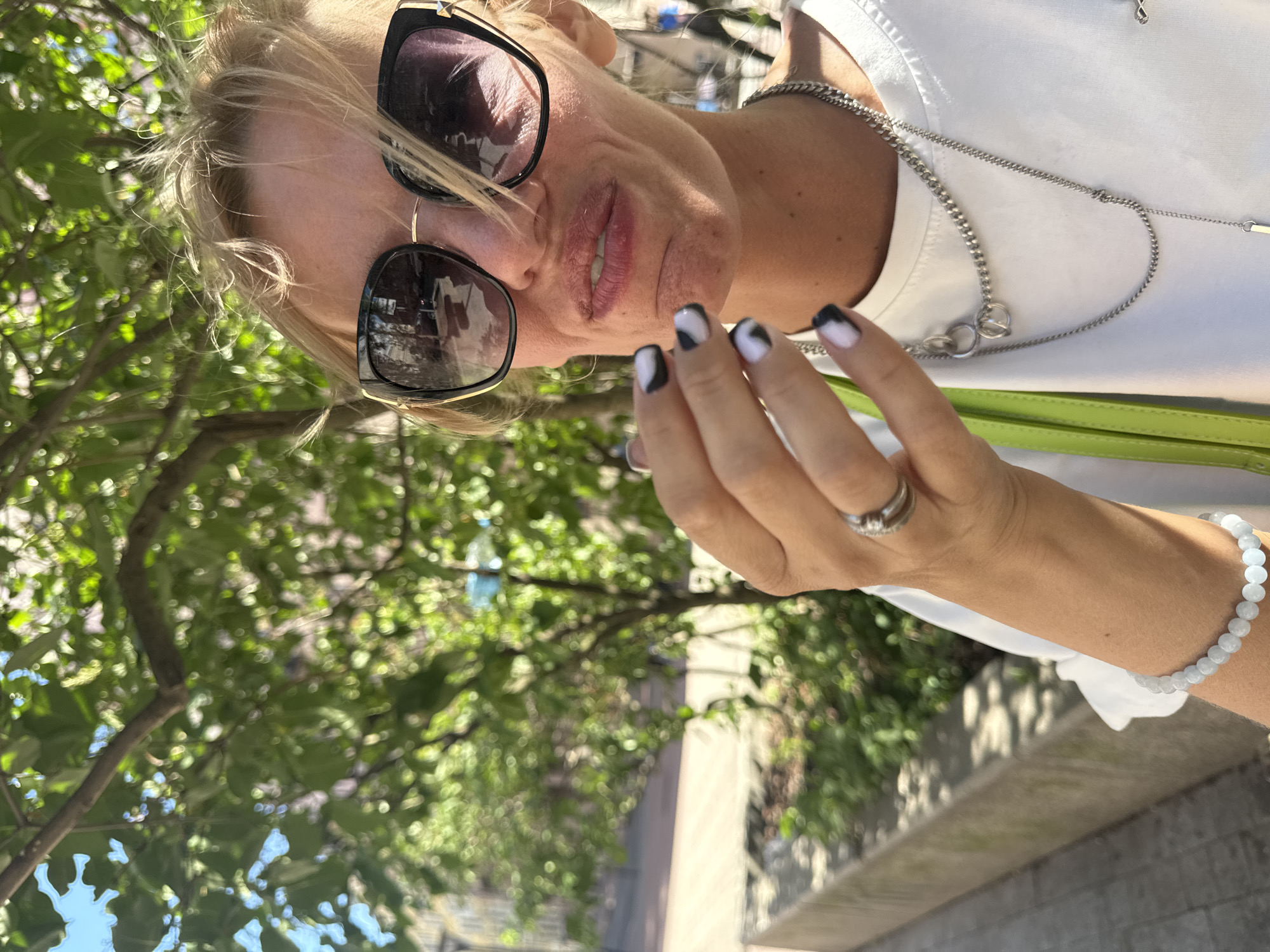In Saint Petersburg, Russia, a woman with blonde hair and sunglasses is captured in a close-up, outdoor shot. She is wearing a white t-shirt and a lime green bag strap is visible. Her hand, with black and white nail art, is near her face. She is also wearing a ring on her left hand and a silver necklace. The background features trees with green leaves and a blurred urban setting.