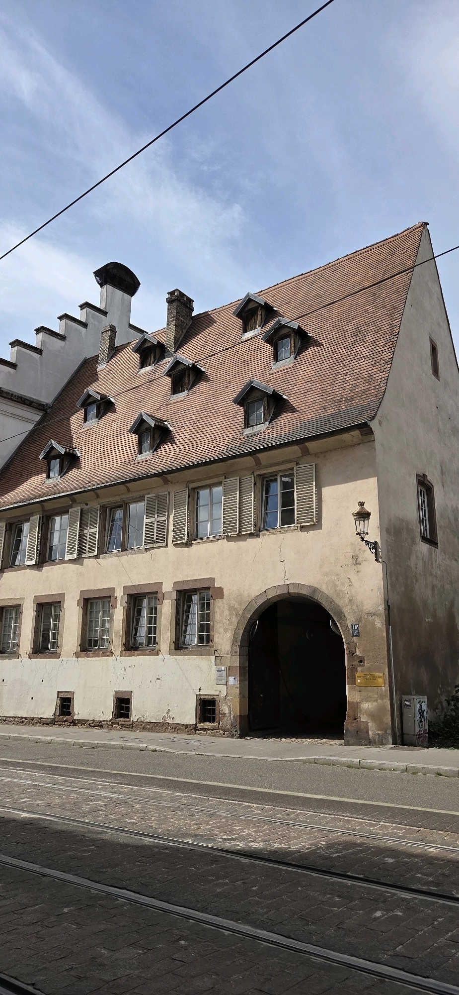 This is a full-frame, eye-level, outdoor, natural light photograph of a section of a building in Strasbourg, France. The building is a historic, multi-story structure with a prominent red tile mansard roof punctuated by numerous dormer windows. The facade is a muted yellow color with visible signs of aging and some cracks. There is a large arched entryway that leads into the interior of the building, suggesting it may have once served as a stable or carriage house. Several windows with wooden shutters are visible on the main facade. A street with tram tracks runs in front of the building. A traditional street lamp is mounted on the exterior wall. The sky above is partly cloudy. The image depicts a street view of a building in Strasbourg's historic quarter, likely along a route served by public transportation.
