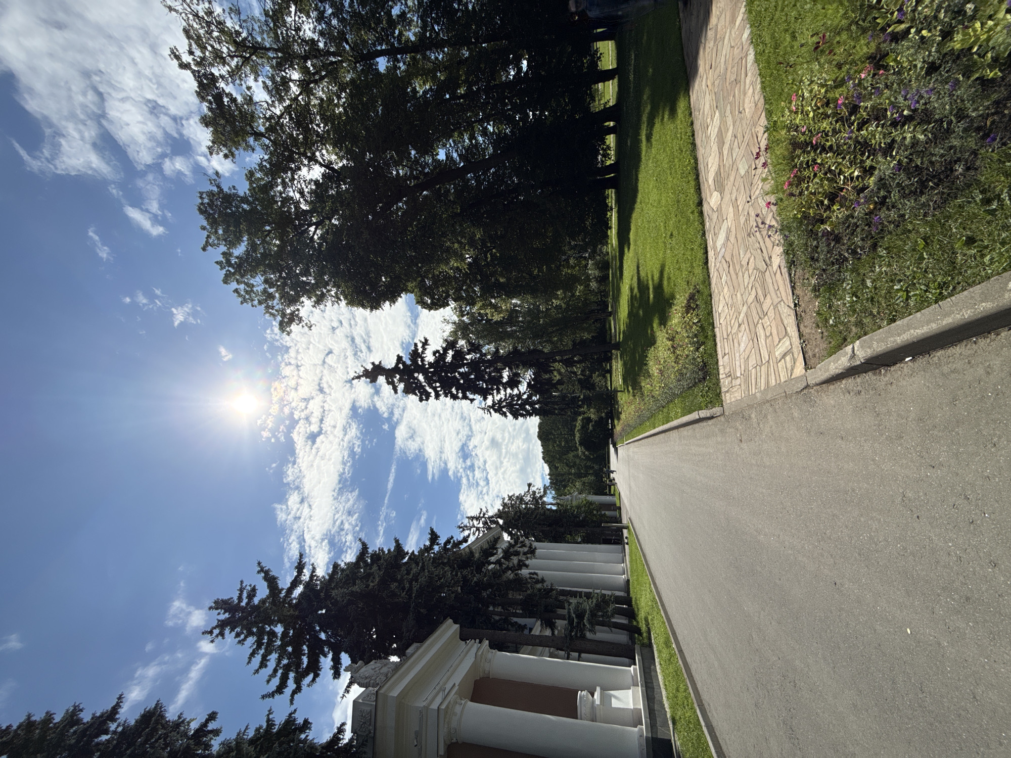 The image captures a sunny day in Moscow, Russia, specifically at the VDNKh (Exhibition of Achievements of National Economy) park. A paved walkway stretches into the distance, flanked by lush green lawns and tall trees. On the left, a classical white building with large columns stands prominently. The sky is a vibrant blue with scattered white clouds, and the sun shines brightly, casting shadows. In the distance, a few people can be seen walking, and some trees create a dense forest on the horizon. The foreground features a flowerbed with blooming pink and purple flowers. The scene conveys a sense of tranquility and natural beauty within an urban park setting.