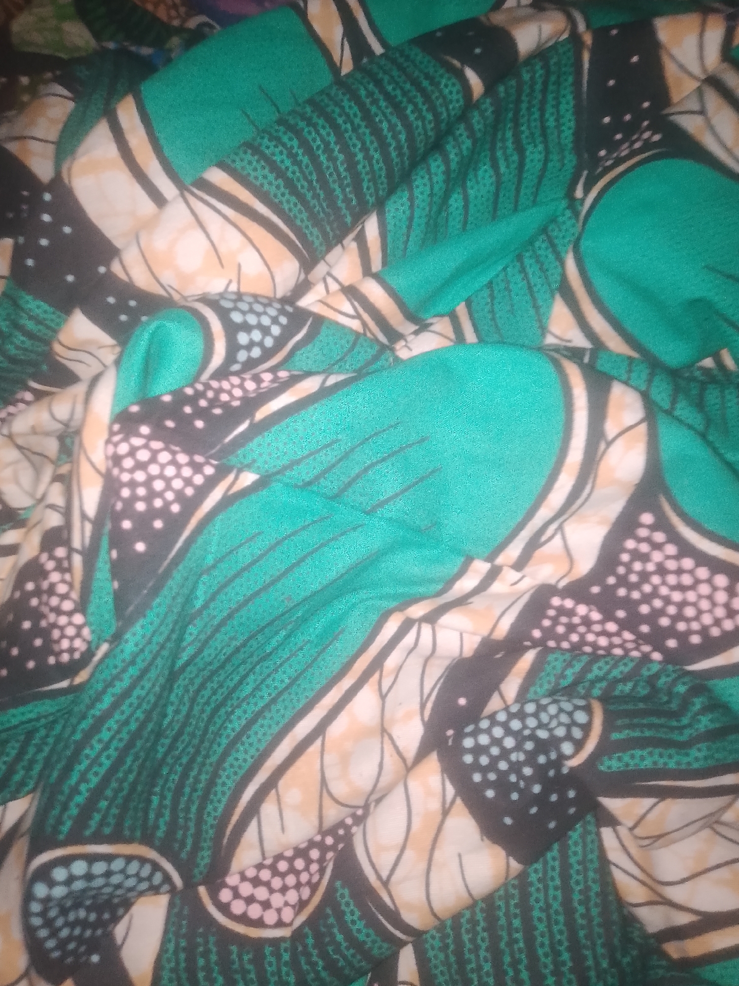 The image displays a close-up, top-down view of a piece of fabric with a bold, abstract pattern. The fabric is predominantly green, with large curved shapes in black and cream, accented by small dots in pink and light blue. The pattern appears to be an African print, characterized by its vibrant colors and geometric or stylized motifs. The fabric is folded and creased, creating shadows and highlights that emphasize its texture and the way it lies. There are no people, objects, actions, or interactions visible in the image. The environment is not discernible from the provided view of the fabric.