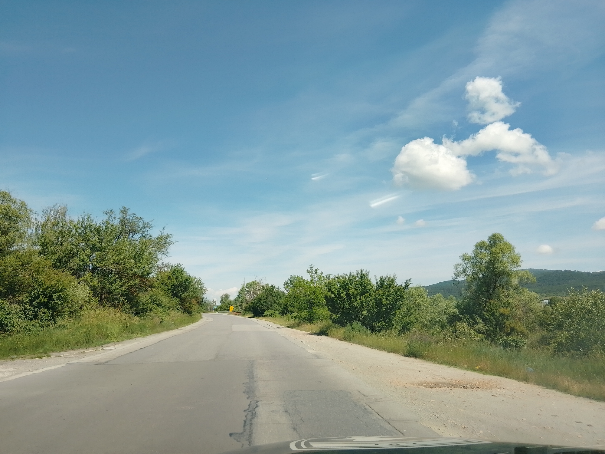 The image shows a view from a vehicle traveling on a paved road through a rural, natural environment. The road appears to be two lanes, with a worn asphalt surface and patchy repairs. The road curves slightly to the left, leading the viewer's eye towards the horizon. On either side of the road, dense green foliage, including trees and bushes, creates a natural border. The ground alongside the road is a mixture of dirt and overgrown grass. In the background, rolling hills covered in vegetation are visible under a bright blue sky with scattered white clouds. The overall scene suggests a daytime, likely sunny, excursion through a sparsely populated, verdant area. There are no people or specific structures identifiable in the image.