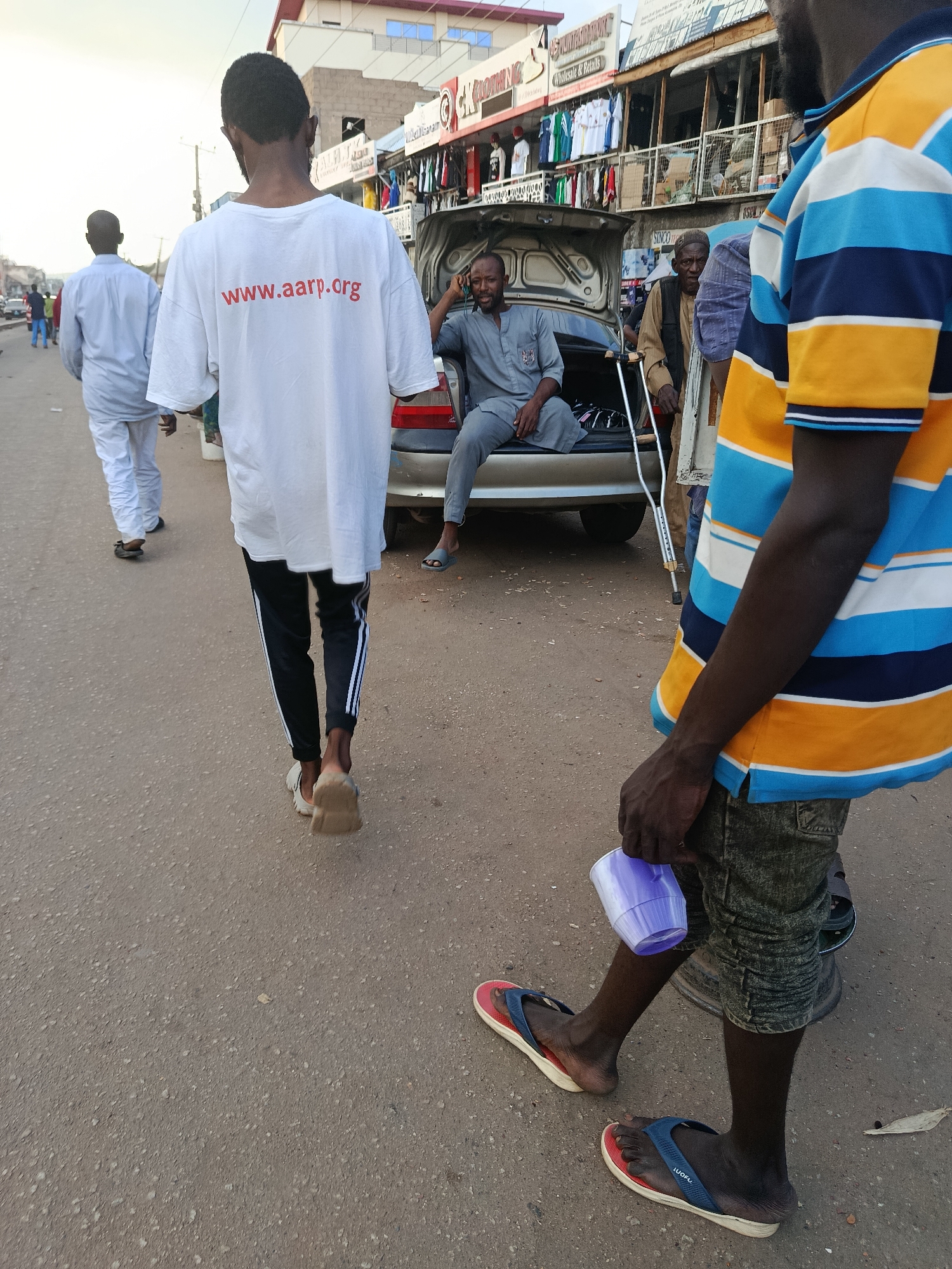 This media file depicts a street scene in Jos, Nigeria. The primary subjects are several men, some walking and others stationary. In the foreground, a person wearing a colorful striped t-shirt and dark shorts walks away from the camera, holding a purple cup. Behind them, another person in a white t-shirt with red text walks down the street. Further back, a man in a gray outfit is seated in the open trunk of a car. Another man, wearing a brown vest and cap, stands near the car, leaning on a crutch. The scene appears to be taking place during the daytime, possibly late afternoon, given the warm lighting. The setting is a paved street lined with shops displaying clothing. Visible text on the white t-shirt reads "www.aarp.org". The overall atmosphere appears casual, with people going about their day on a street.