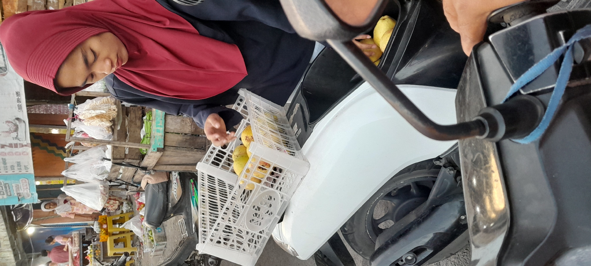 In Rogojampi, Indonesia, a young woman wearing a red hijab and a dark blue garment is standing next to a white motorcycle. She is reaching into a white plastic crate filled with yellow fruits, likely guavas, that is placed on the motorcycle's seat. More of the same fruit is visible in the storage compartment of the motorcycle. In the background, a market stall is visible with various goods, including bagged snacks and produce, with other individuals present. The scene depicts a common everyday activity at a local market.