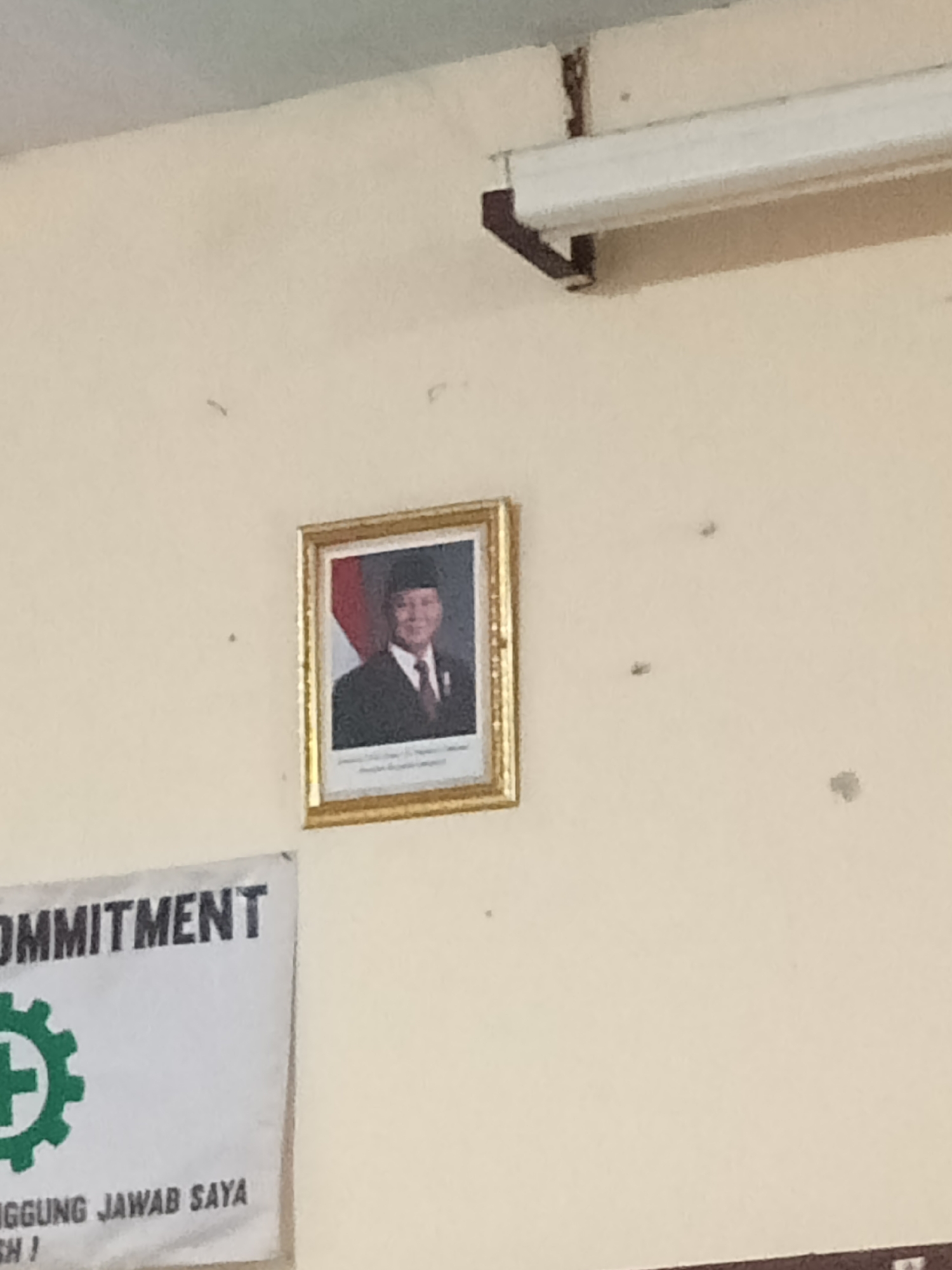 An official portrait of a man in a dark suit, white shirt, and red tie, wearing a traditional black cap, is displayed in a gold frame on a pale yellow wall. To the left of the portrait, a white banner with black text reading "OMMITMENT" is partially visible. Below the text, a green circular logo with a white cross and gear-like spokes is prominent. Further down the banner, more black text is visible, including "NGGUNG JAWAB SAYA" and "SH!". A fluorescent light fixture is mounted above and to the right of the portrait. The image appears to be taken indoors, possibly in a public building or office, in Secang, Indonesia. The lighting is artificial and casts faint shadows.