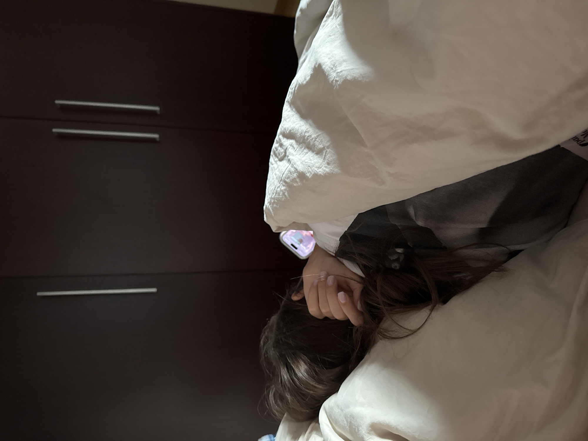 A person is in bed, partially covered by a white blanket. The person is holding a phone in their hand. To the left of the bed is a dark-colored cabinet with silver handles. The location is Unknown, Unknown.
