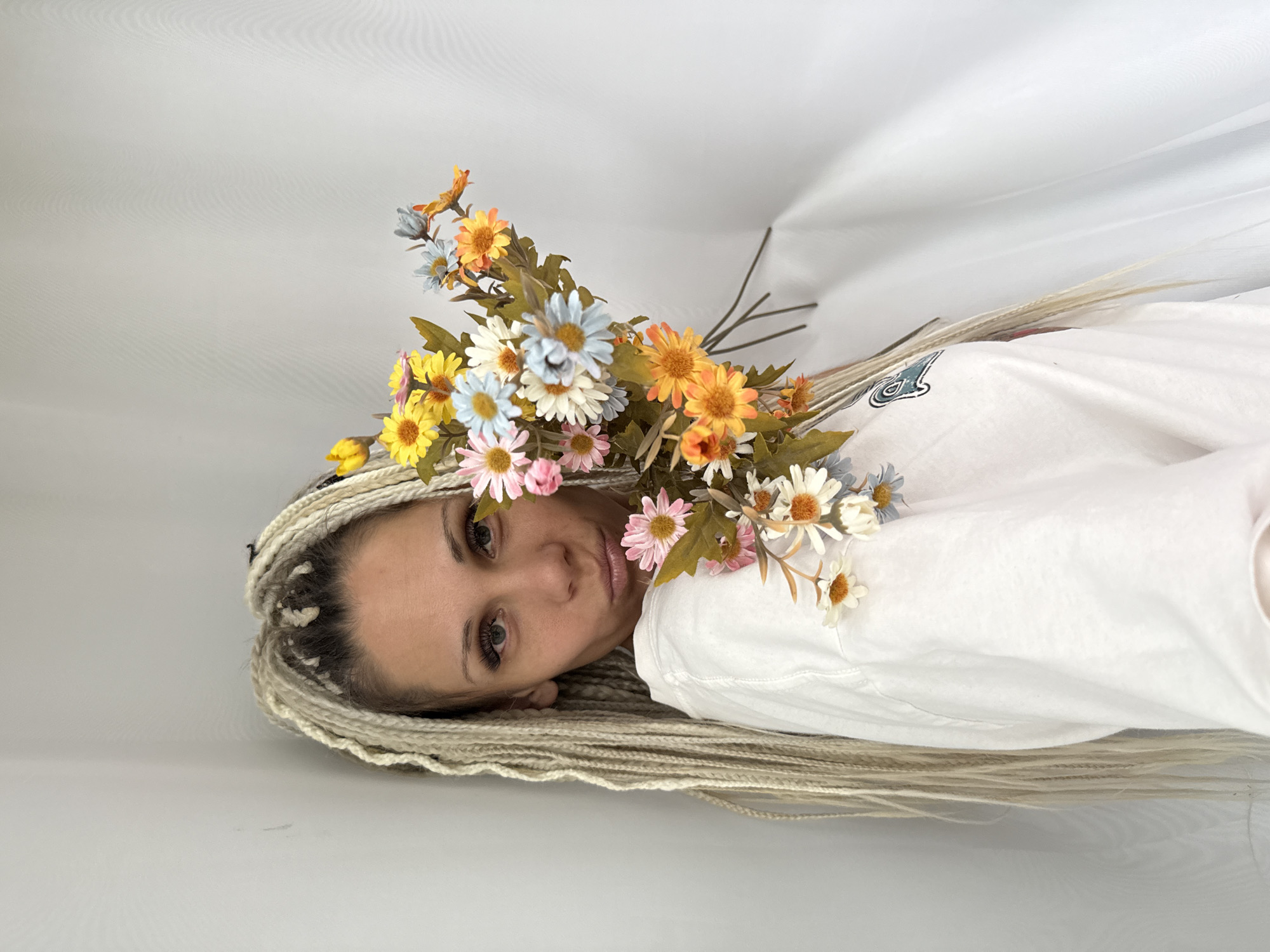 The image shows a woman with long, blonde braids holding a bouquet of artificial daisies. She is wearing a white t-shirt. The background is a white curtain and a light grey wall. The image does not contain any information about the location in the city of Rusanovo, Russia.