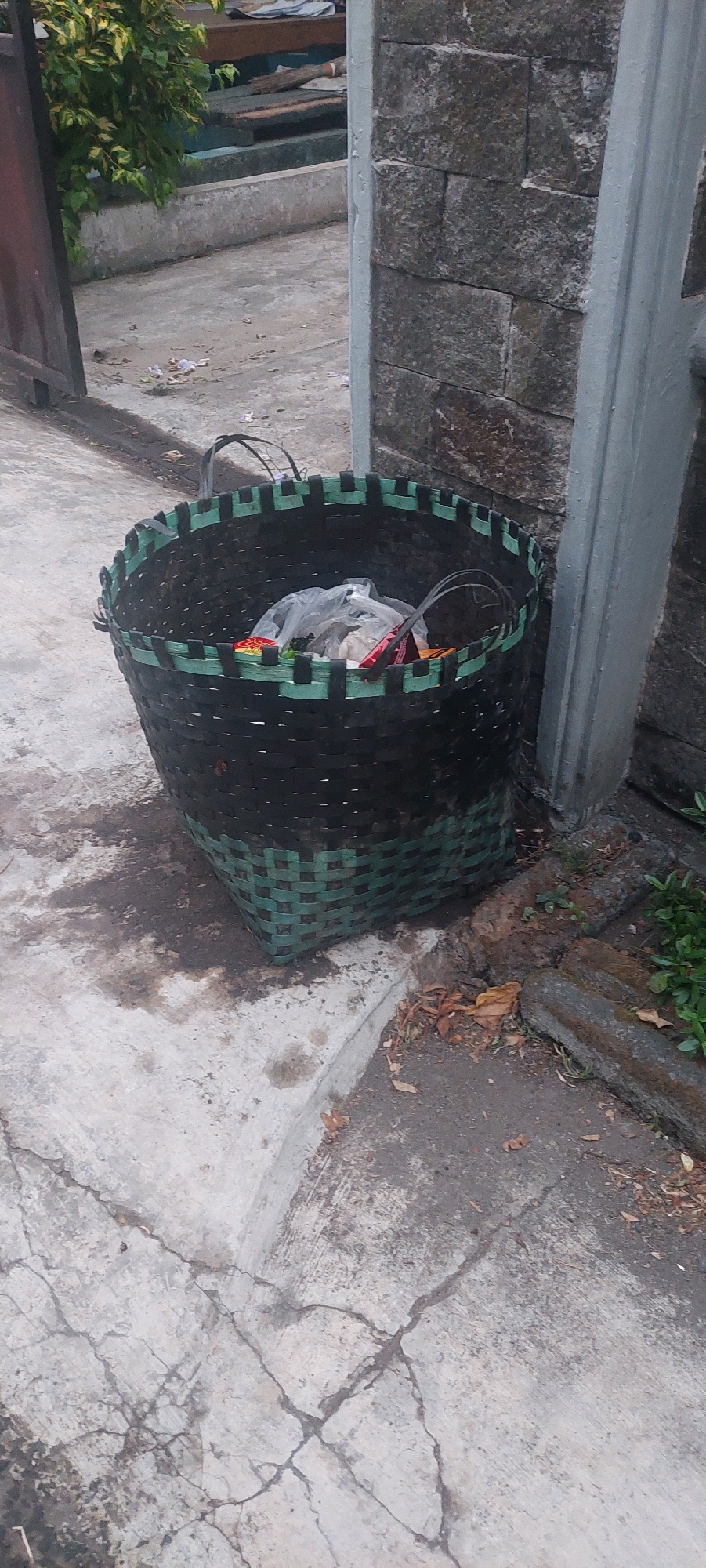 A woven basket, predominantly black with green accents, sits on a cracked concrete ground adjacent to a stone wall and a grey concrete pillar. The basket contains several plastic bags filled with debris, including what appears to be food wrappers and other refuse. The surrounding environment consists of a pathway and some low-lying vegetation. The scene is outdoors, likely in an urban or suburban setting within Sukodono, Indonesia, given the visual cues such as the style of construction and vegetation. There are no people present in the image.