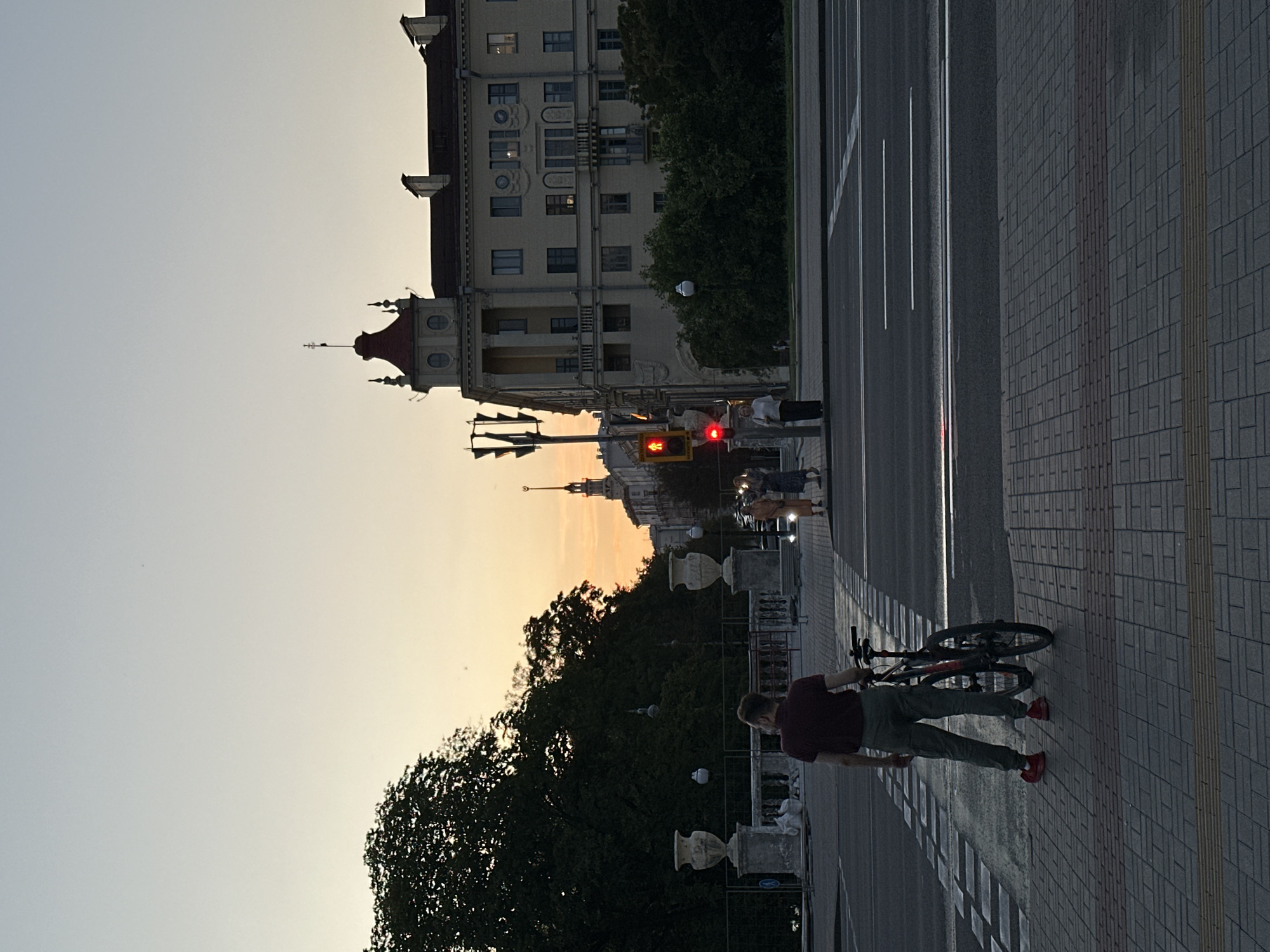 A silhouetted man stands on a sidewalk with a bicycle at a crosswalk, facing away from the camera. He is wearing a dark short-sleeved shirt and light-colored pants. The bicycle is dark-colored with red accents. The crosswalk is marked with white lines on the asphalt. In the background, a red traffic light is illuminated, and two women are standing near a traffic pole. Across the street, a large, ornate building with a red-roofed tower is visible, along with trees and the cityscape extending into the distance. The sky is a gradient of soft orange and pale blue, indicating dusk.