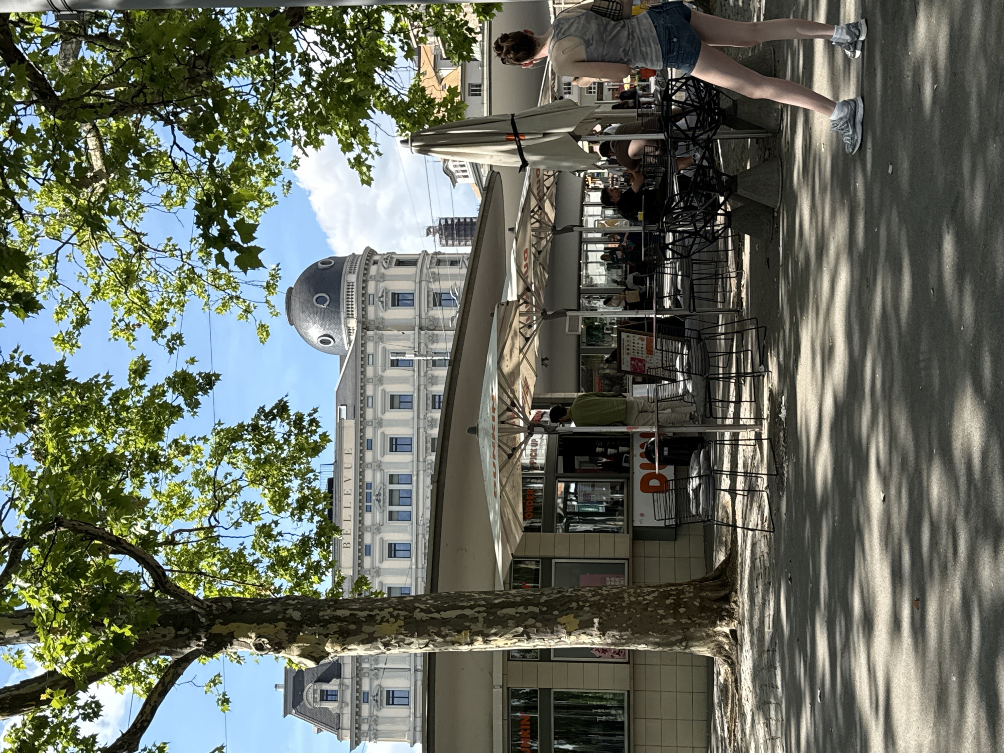 This image depicts an outdoor seating area of a cafe, likely a Dunkin' Donuts given the visible signage, located in Zurich, Switzerland. The cafe is situated in front of a large, ornate building with the word "BELLEVUE" emblazoned across its facade. The building features classical architecture with a domed roof. In the foreground, a woman with dark hair pulled back is standing, looking towards the cafe. She is wearing a camouflage tank top and denim shorts, with white socks and grey athletic shoes. The cafe has several tables and chairs set up under large umbrellas. In the background, other patrons are seated at tables. A large tree with speckled bark is prominently featured on the left side of the frame, casting shadows on the paved ground. The sky is blue with scattered white clouds.
