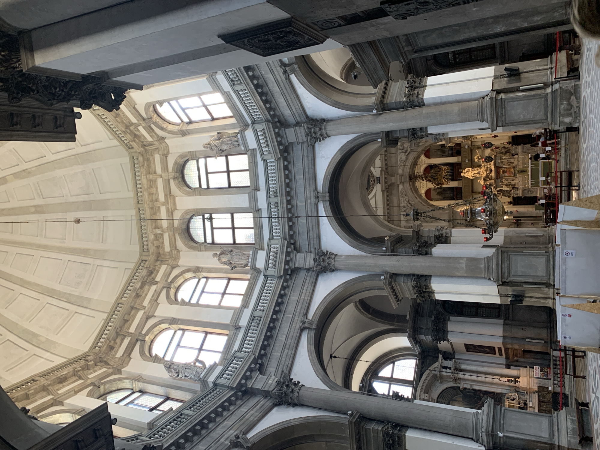 This is an interior photograph of the Basilica di Santa Maria della Salute in Venice, Italy. The viewpoint is from the nave, looking towards the altar. The image captures the grand scale of the interior, dominated by a large dome and intricate architectural details.
Key elements visible include:
* **Architecture:** A prominent dome with coffered panels stretches upwards. A circular balcony with balustrades runs around the base of the dome, supported by large, ornate columns and arched openings. There are multiple levels of arched windows, some with stained glass, admitting natural light. Sculptures of figures are placed on pedestals along the upper levels. The lower sections of the church feature large, fluted columns and more arched doorways.
* **Altar and Apse:** In the background, the altar area is visible, adorned with statues and religious iconography. A large pipe organ is situated above the altar.
* **Lighting:** Natural light streams in through the windows, illuminating the space. Chandeliers hang from the ceiling, though they appear unlit.
* **Furnishings:** Benches are visible in the nave. Several large white panels, possibly protective coverings or temporary exhibits, are placed in the foreground.
* **People:** A few individuals are present in the distance near the altar, appearing to be tourists or visitors. One person's hair is visible in the lower right corner of the frame, suggesting the photographer is among others.
* **Atmosphere:** The overall impression is one of solemnity and grandeur, characteristic of a large Baroque church. The stone and marble construction contribute to a sense of permanence and historical weight.
The photograph is taken from a low angle, emphasizing the height and volume of the interior. The lighting creates a contrast between bright, sunlit areas and shadowed regions.