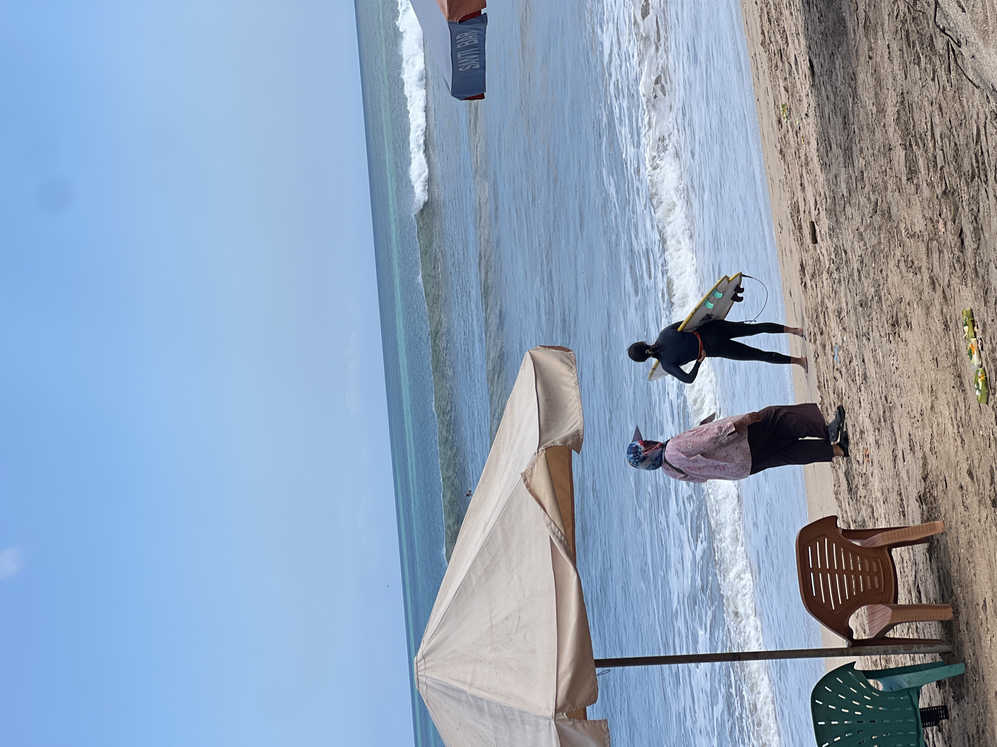 An outdoor scene on a sandy beach in Kuta, Indonesia, with the ocean and sky dominating the background. A large, tan beach umbrella is in the foreground, casting a shadow. To its right, two individuals are standing on the sand. One is a person wearing a black wetsuit and holding a yellow surfboard. The other is a person wearing a pink patterned top and dark pants, with a patterned head covering. Two plastic chairs, one green and one brown, are visible in the lower left corner. Further out in the ocean, waves are breaking, and at least two people appear to be in the water, possibly surfing. Another umbrella with the text "SANTI BAR" is visible in the mid-right distance. The sand is light brown and appears slightly damp in places. The sky is a clear blue with a faint cloud.