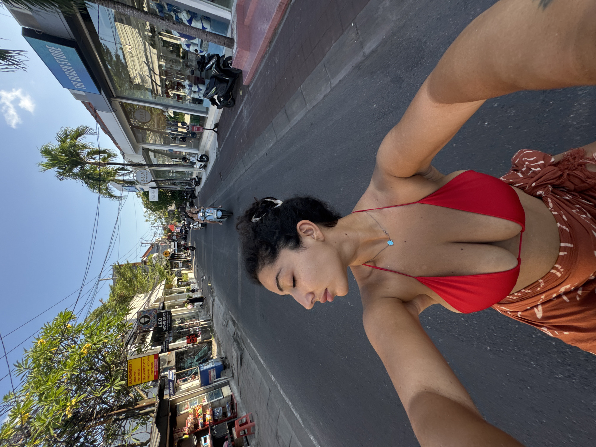 A woman in a red bikini top is on the streets of North Kuta, Indonesia. She is likely taking a selfie with her arm outstretched. Businesses line the street with visible signs like "THE BEACH STORE" and "IKA HOMESTAY." Motorbikes are parked and moving along the roadway, which is surrounded by trees and tropical vegetation. The sky is clear.