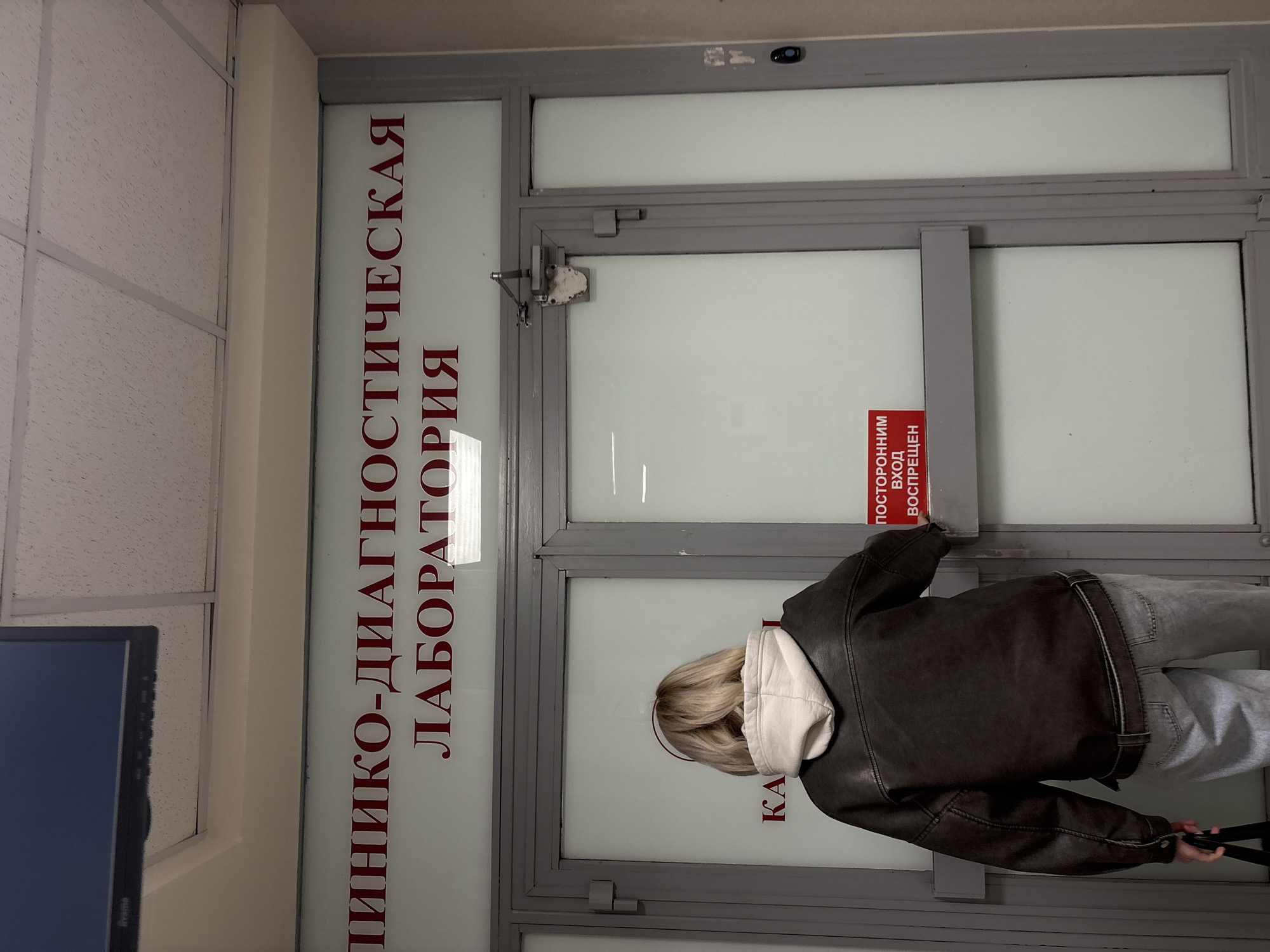 The image shows a person standing in front of a door with a sign in Russian that reads "КЛИНИКО-ДИАГНОСТИЧЕСКАЯ ЛАБОРАТОРИЯ," indicating a clinical diagnostic laboratory. A red sign on the door reads "ПОСТОРОННИМ ВХОД ВОСПРЕЩЕН", which translates to "Entrance forbidden to unauthorized persons." The person is wearing a dark brown leather jacket with a white hoodie underneath and light gray pants. They are reaching for the door handle. The door is framed in gray metal and has frosted glass panels. A monitor is visible in the lower-left corner. The location is Saint Petersburg, Russia, based on the Russian text.