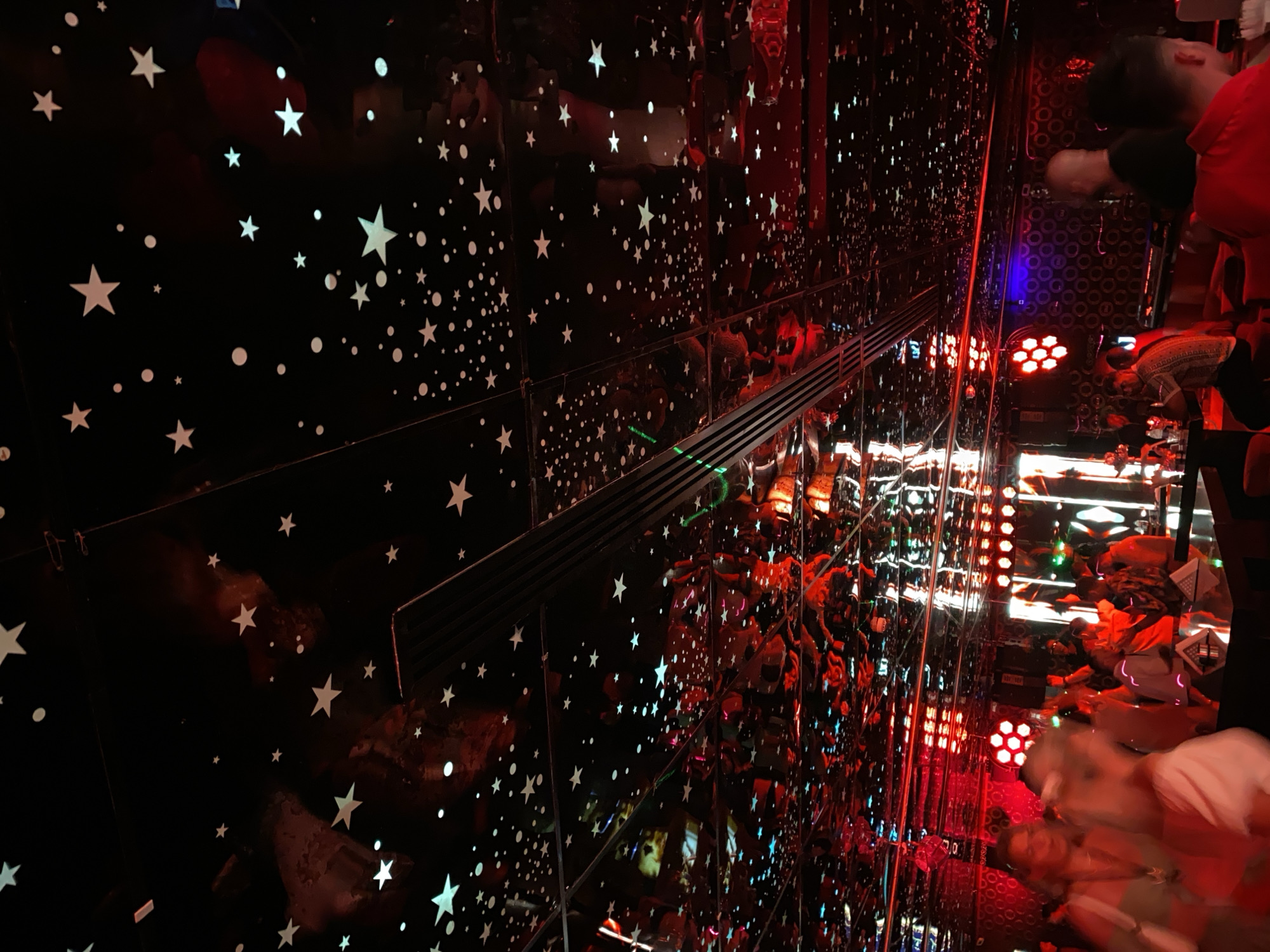 This image shows the interior of a nightclub, likely in Sharm El Sheikh, Egypt, with a distinctive starry ceiling. The ceiling is black and covered in numerous white stars and dots, creating a celestial effect. The room is dimly lit, with strong red and blue lighting illuminating the space. Reflections from the mirrored ceiling show more of the starry pattern and the red lights. In the foreground and middle ground, several people are visible. Some are blurred due to movement, suggesting they are dancing or mingling. Others are more defined, appearing to be standing or sitting at tables. The overall atmosphere appears lively and energetic, typical of a nightclub setting. There are tables with napkins, bar counters, and stage lighting visible in the background.