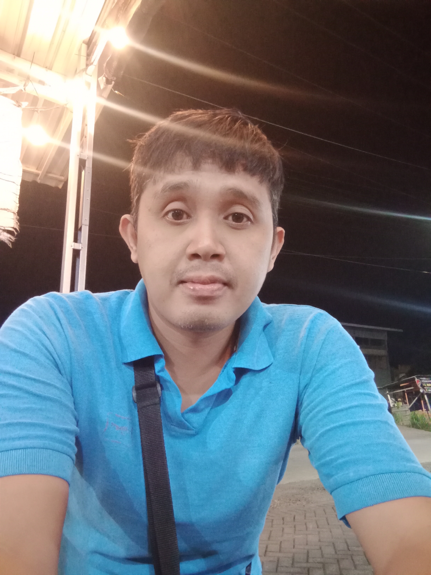 The image presents a close-up, frontal portrait of an adult male of apparent Southeast Asian ethnicity, estimated to be in his late 30s to early 50s, captured in the city of Cangkring, Indonesia. He has short, dark brown hair with a fringe across his forehead, and visible stubble around his mouth and chin. His dark eyes are open and directed straight at the viewer, maintaining a neutral facial expression with slightly pursed lips.
He is dressed in a light blue, short-sleeved polo shirt with a collar, the top two buttons unfastened. A black fabric strap, possibly from a bag or camera, is visible diagonally crossing his chest from his right shoulder.
The scene is set at night, indicated by the dark background, with prominent artificial lighting. In the upper left background, bright, warm yellow light sources, possibly bare bulbs or string lights, are visible under a light-colored, corrugated metal roof structure, creating significant lens flare. The upper right and mid-right background are mostly dark, with faint horizontal light streaks suggesting distant light sources or motion blur. The lower portion of the image reveals an outdoor ground surface paved with light-colored, rectangular paver blocks. The environment suggests an outdoor or semi-outdoor commercial or public area.