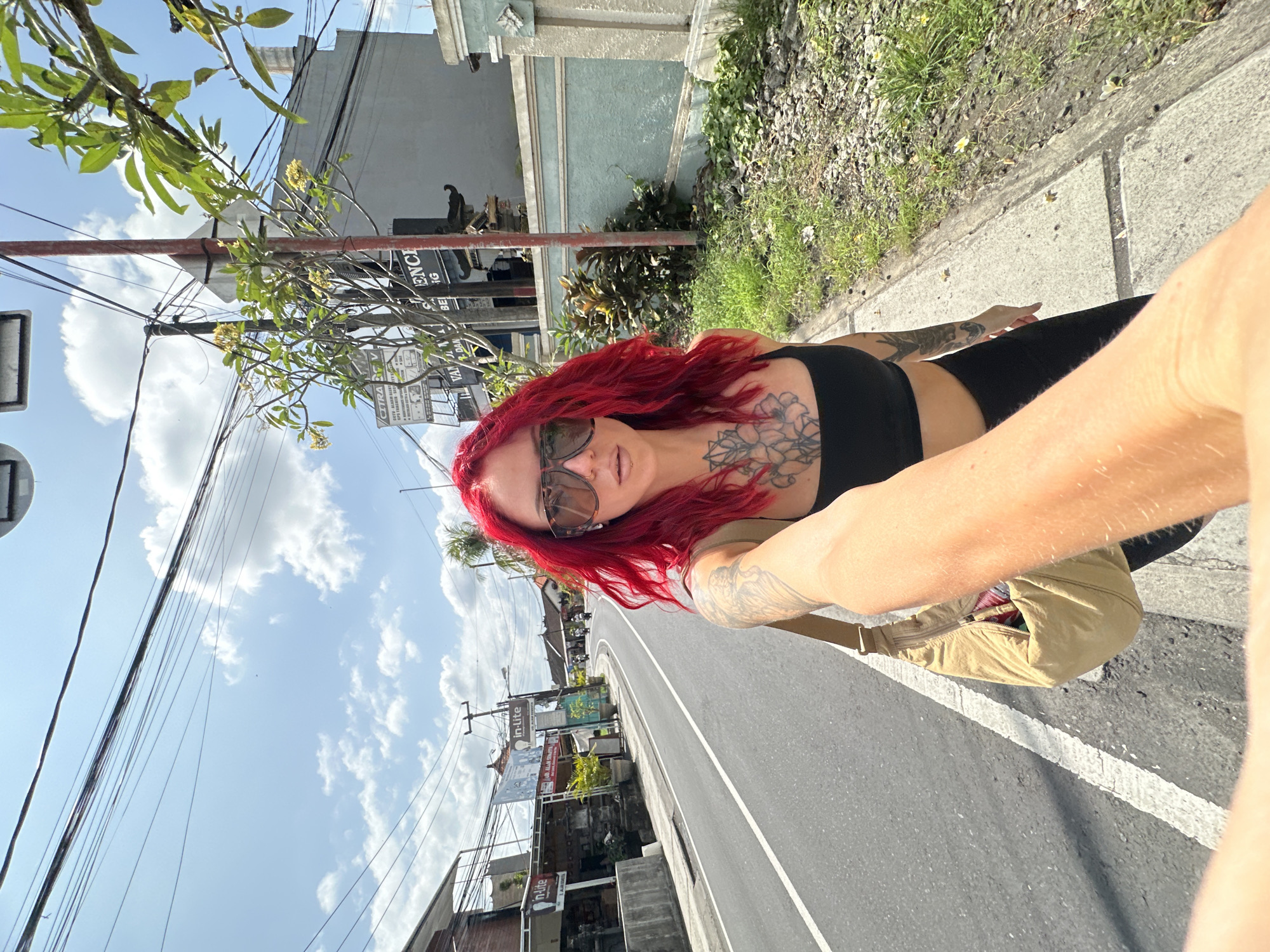 A female with red hair, sunglasses, and tattoos is taking a selfie in North Kuta, Indonesia. She's wearing a black top and black pants. The background includes a street, buildings with signs (one reading "in-lite"), trees, and a sky with clouds. She has a light brown bag around her waist.
