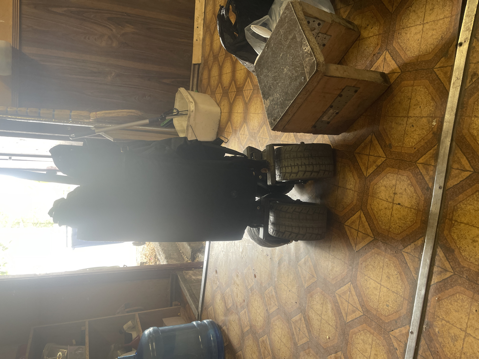 The image depicts an indoor utility area or workshop, in the city of Unknown, Unknown, with an open doorway leading outdoors. A large, dark-colored, motorized object with wide, knobby tires occupies the center foreground. To the left, a blue five-gallon water bottle rests on a worn hexagonal-patterned linoleum floor. Further left, a light-colored wooden shelving unit contains various containers. The open doorway reveals concrete steps and a bright outdoor scene with a partial view of a blue trash bin and foliage. To the right of the central object, several brooms and a dustpan lean against a dark wood-paneled wall. A white plastic bucket sits on the floor nearby. In the immediate right foreground, a homemade wooden step stool or block with a dark, textured top and metal reinforcements is present. A black trash bag is partially visible behind the stool. A silver metal strip runs horizontally across the bottom edge of the frame. The area is illuminated by strong natural light entering from the doorway. No individuals are present in the scene.