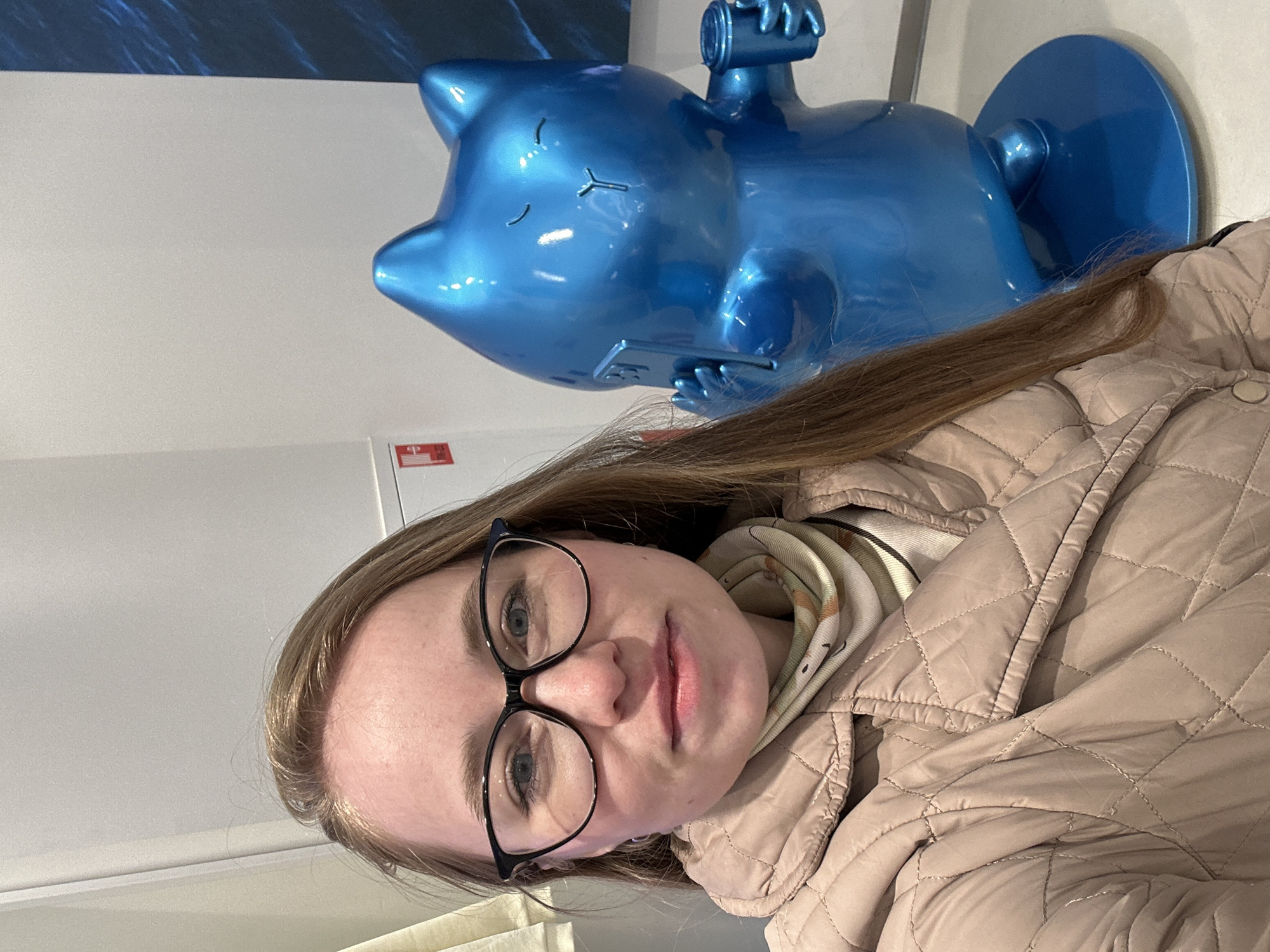 The image, taken indoors and rotated 90 degrees clockwise, captures a young woman posing in front of a unique blue statue. The woman, positioned on the right side of the frame (when correctly oriented), has light skin, long brown hair, and wears black-framed glasses. She is dressed in a light beige quilted jacket over a patterned scarf or hooded top, looking directly at the camera with a neutral expression.
Behind her, on the left, stands a large, glossy metallic blue sculpture of a stylized cat-like creature. The creature is depicted lying on its back, holding a small bottle or can above its head with one paw and a smartphone on its belly with the other. Its eyes are closed, conveying a peaceful and relaxed state.
The background consists mainly of a clean, plain white wall, with a darker blue element visible at the top, possibly part of a display or another artistic feature. A notable detail is a small red and white rectangular sign on the white wall behind the statue, which clearly displays the Chinese characters "中国邮政" (Zhōngguó Yóuzhèng), meaning "China Post," along with a symbol and what appears to be a barcode or QR code. The setting appears to be a public indoor space, such as an exhibition, a shop, or a cultural center, located in Saint Petersburg, Russia, as suggested by the visible text and the provided location context. The lighting is bright and even, typical of an indoor environment.