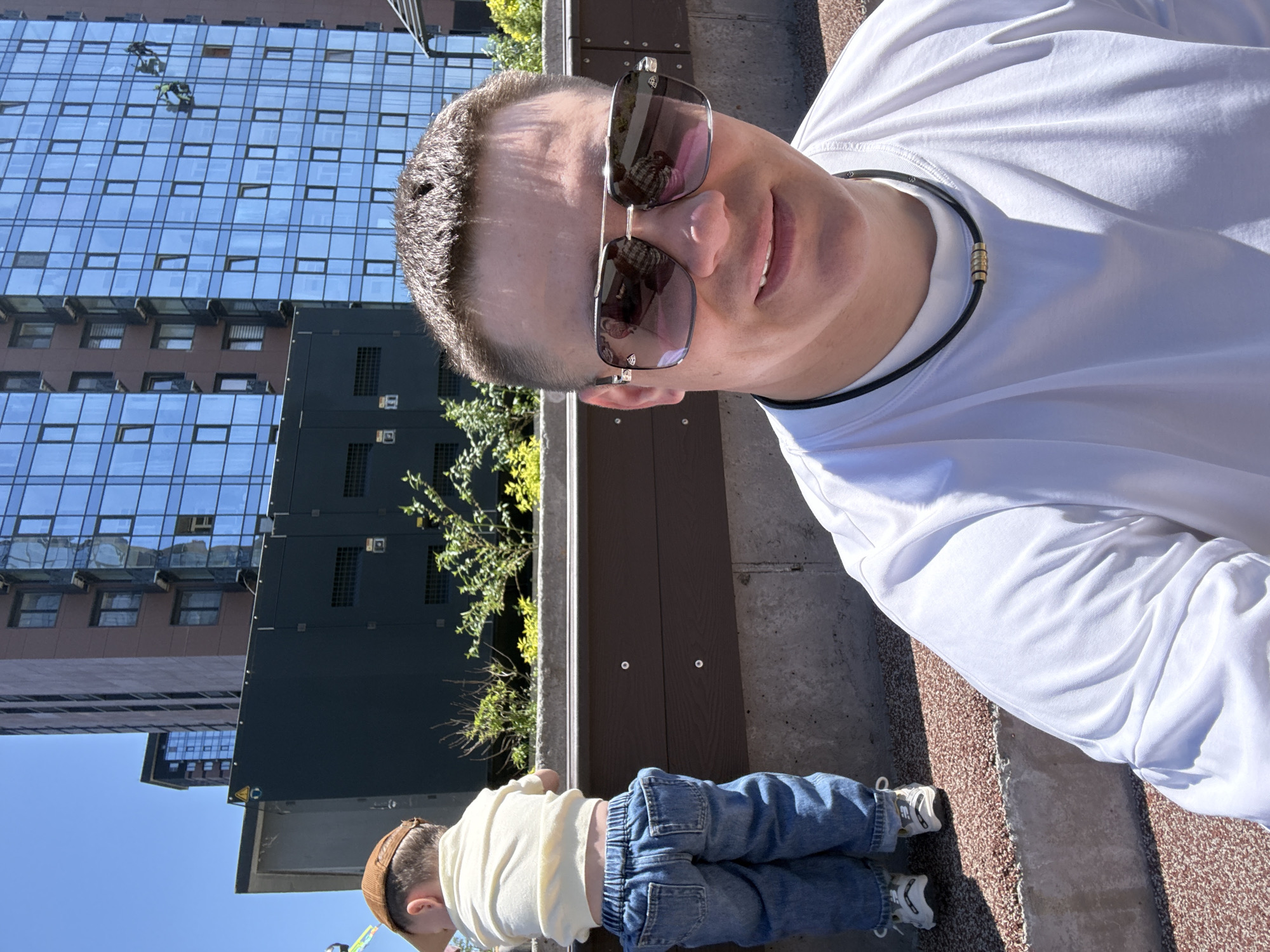 A man is taking a selfie in front of a building in Vladivostok, Russia. He is wearing sunglasses and a white shirt. A child is lying on the ground near a low wall, also facing the building. The man is looking at the camera and smiling. The building in the background is made of glass and has many windows. Window washers appear in the reflection of the glass windows on the building. The day appears sunny.