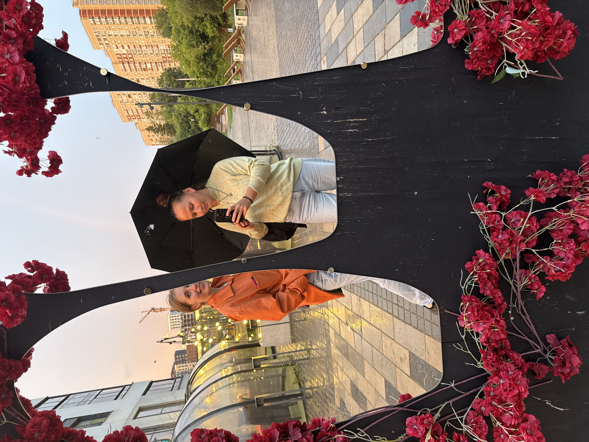 The image is a reflection in a curved, black-bordered mirror, showing two women and an urban scene. The mirror is adorned with artificial red flowers. The scene is a public outdoor space in Izhevsk, Russia, likely a plaza or promenade, as indicated by the paving stones and modern buildings.
In the upper reflection, a woman with her hair in a bun is holding a black umbrella and a smartphone, appearing to take a selfie. She is wearing a light yellow sweater and light blue jeans. A building with many windows and balconies is visible in the background, along with trees and a clear sky suggesting it is daytime, possibly late afternoon given the warm light.
In the lower reflection, another woman is smiling at the camera. She is wearing an orange quilted jacket and light blue jeans. Behind her, there is a glass-enclosed structure, possibly a cafe or shop, and strings of lights are visible, hinting at an evening ambiance or a decorative lighting setup. Construction cranes in the distance suggest ongoing development in the city. The sky in this reflection shows a gradient of colors, from light blue to orange, reinforcing the idea of dusk or dawn.
The overall mood is relaxed and cheerful, with both women smiling. The artificial flowers add a decorative and somewhat whimsical touch to the urban setting. There is no visible text in the image.