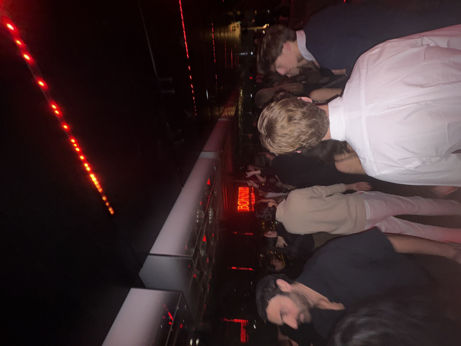 The image depicts a crowded, dimly lit interior scene within the Bonnie Club, located on the rooftop of the SO/ Paris hotel at 10 Rue Agrippa d'Aubigné, in the 4th arrondissement of Paris, France. The environment is dominated by artificial red lighting, including linear LED strips on the dark ceiling and walls. White, reflective rectangular panels are also visible on the ceiling, reflecting the red light.
A prominent red neon sign displaying "BONNIE" in a larger font and "CLUB" beneath it is visible in the mid-background. Numerous individuals are present, populating the space. In the immediate foreground, on the left, a man with dark hair and a beard is seen in profile, wearing a dark collared shirt. Directly in front of the camera, the back of a person's head with light brown, wavy hair is visible, wearing a white collared shirt. To the left of this person, another individual is seen from the back, wearing a light-colored long-sleeved top and light pants. To the right, a person with dark hair and a dark collared top is seen in profile. Further into the background, a dense crowd of people is visible, with some individuals appearing to have raised hands, suggesting a lively social gathering or club activity. The overall scene indicates a nocturnal social event.
