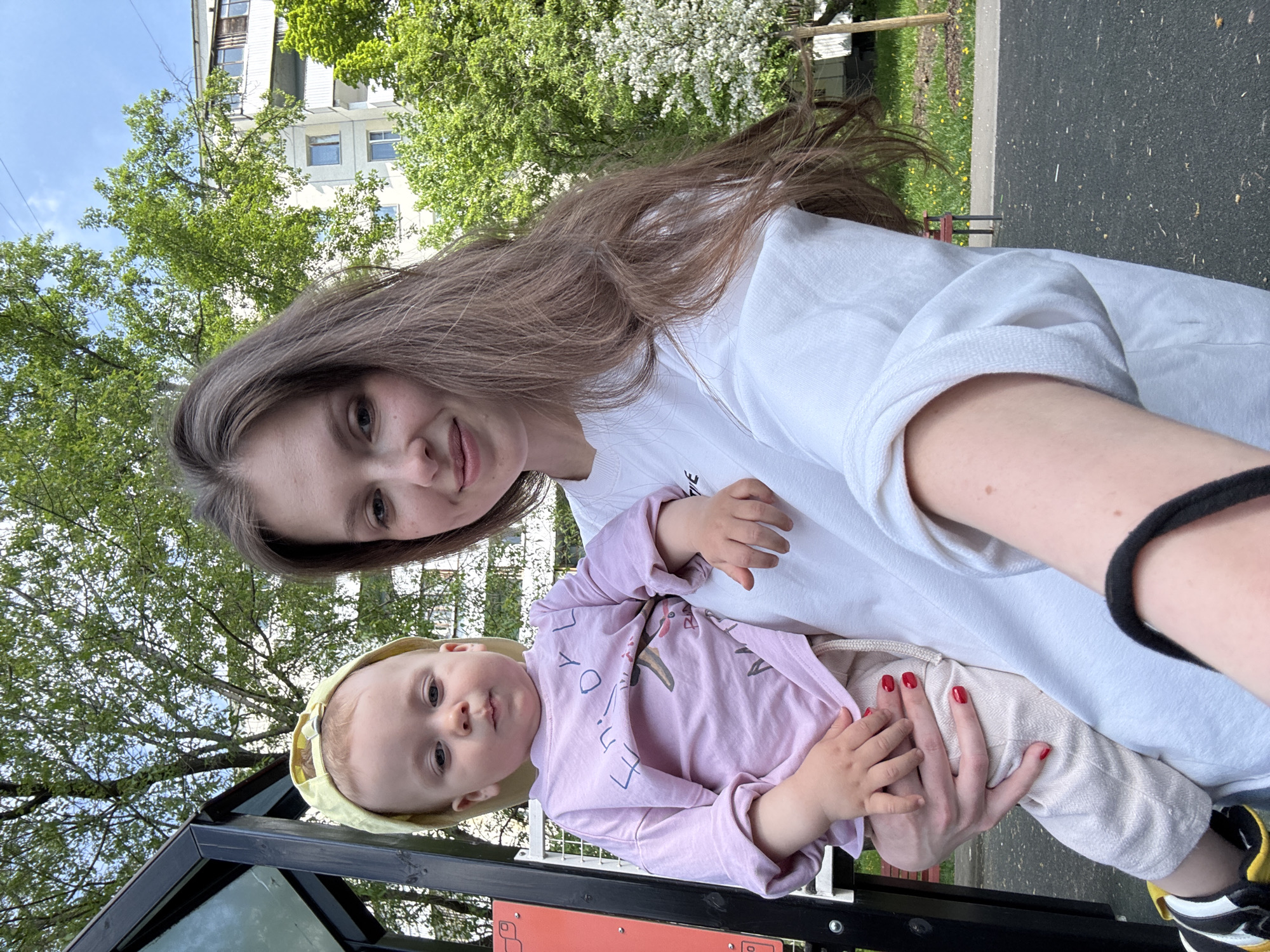 A woman holds a baby in an outdoor setting. The woman has long brown hair and is wearing a white shirt. The baby is wearing a light purple outfit and a yellow hat. The background includes trees, a building with light-colored walls, and what appears to be a playground structure. The environment is consistent with an urban park-like setting. Based on visual details, it is not possible to confirm with certainty the location is Moscow, Russia, but the architectural style of the building could be seen in different parts of Russia.