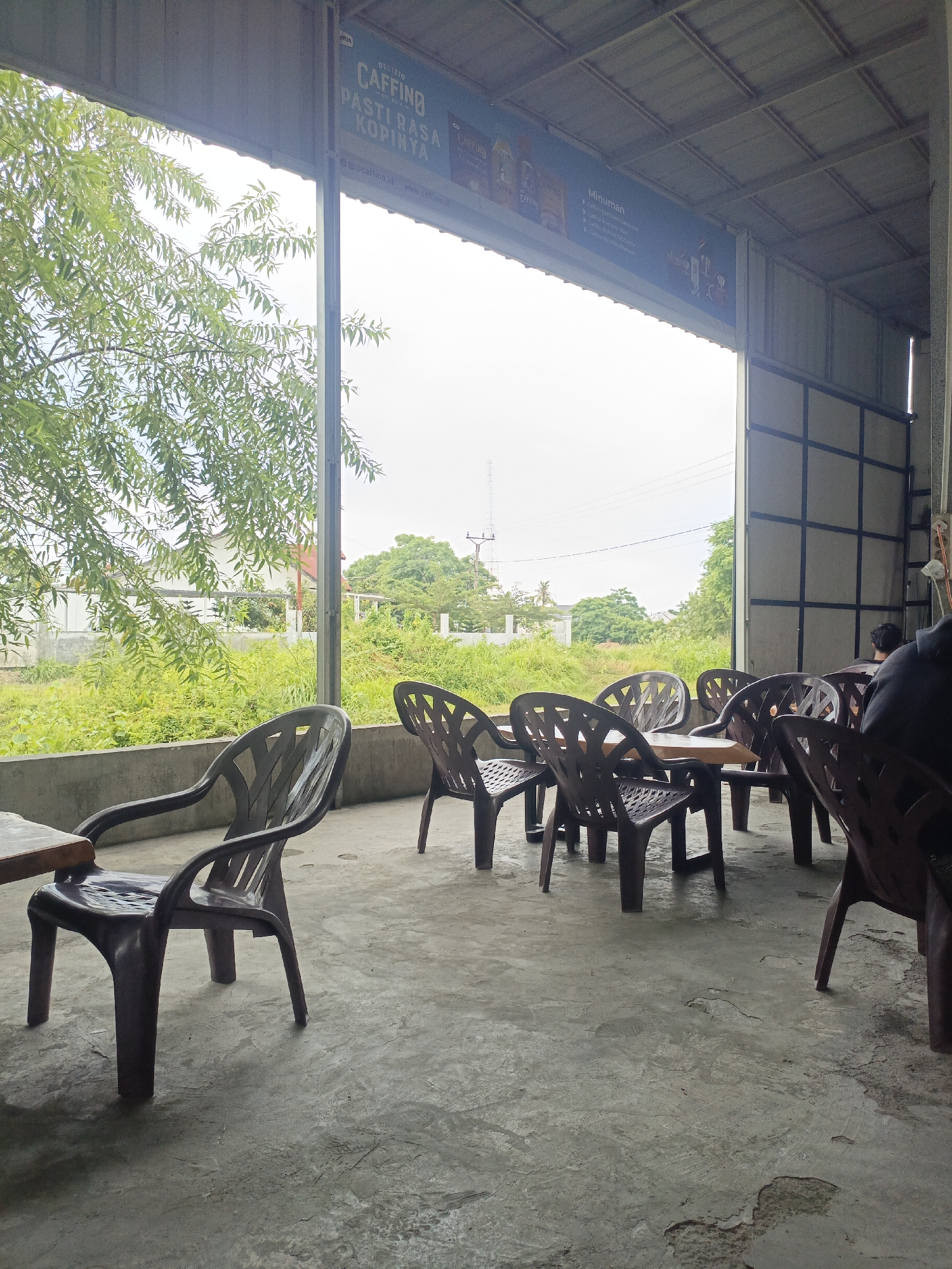 The image depicts an outdoor seating area of a cafe, likely in Lhokseumawe, Indonesia, characterized by its open-air design with a metal roof supported by visible beams. The floor is concrete, showing signs of wear and tear. Numerous plastic chairs, predominantly dark brown, are arranged around tables. In the foreground, a single plastic chair is positioned close to a wooden tabletop, suggesting a solitary diner or a table setting.
Beyond the seating area, lush green vegetation fills the background, with trees and shrubs indicating a possibly rural or suburban outskirts setting. In the distance, a white building with a red roof is visible, and further out, telephone poles and more trees suggest a developed, yet green, landscape. The sky is overcast, contributing to a diffused light.
A banner is visible on the underside of the roof, displaying text related to "CAFFINO" and "MINUMAN," implying the cafe serves beverages. A hint of a person, dressed in dark clothing, is visible in the far right of the image, seated and facing away from the camera, adding a subtle human element to the scene. The overall atmosphere is casual and relaxed, typical of a local eatery or rest stop.