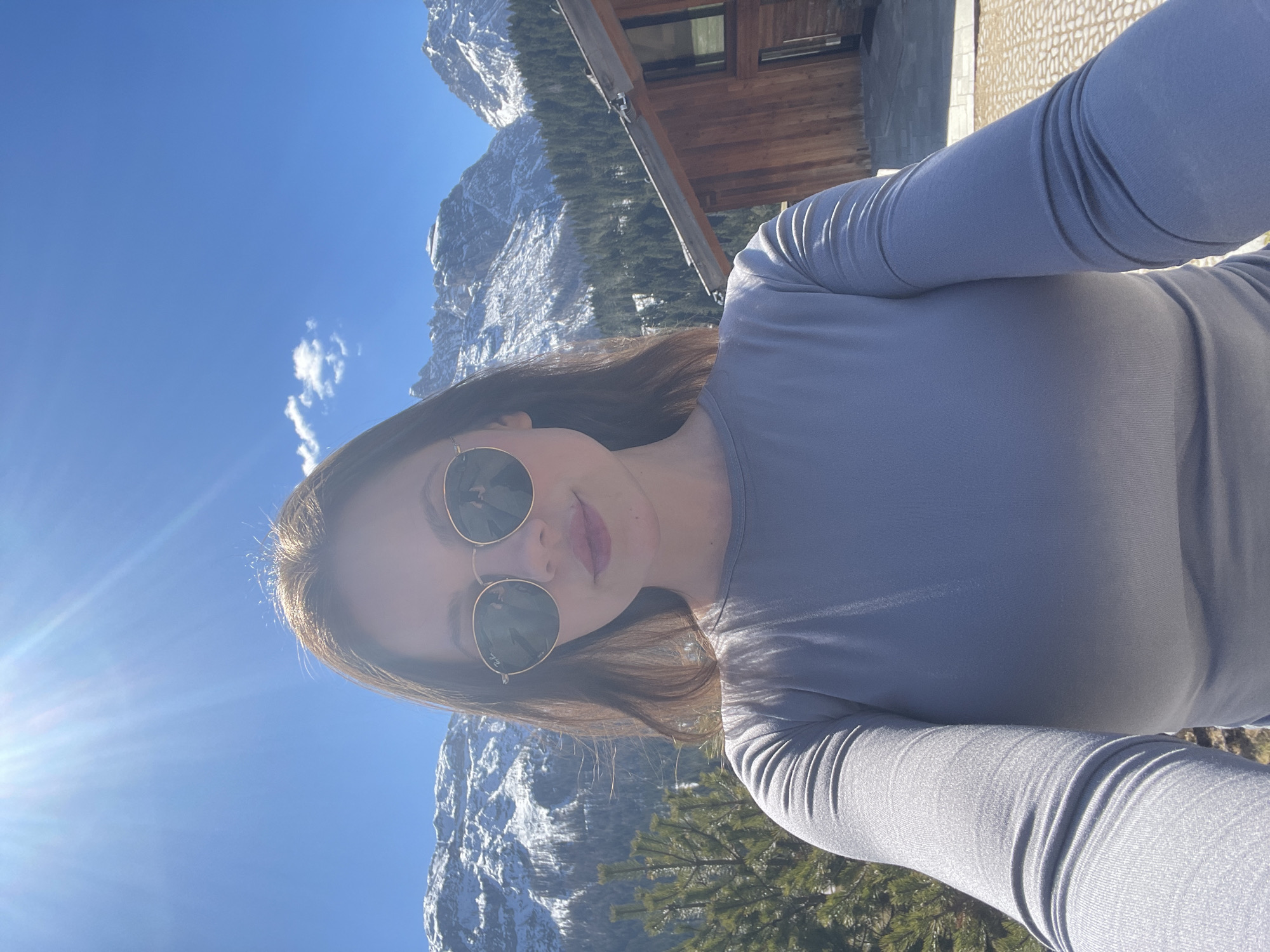 A selfie shows a woman wearing sunglasses, a long-sleeved gray shirt, and standing outside with mountains in the background. Snow is visible on the mountain tops. Some trees and a wooden building are also visible. The photograph was taken in the city of Sappada / Plodn / Sapade, Italy.
