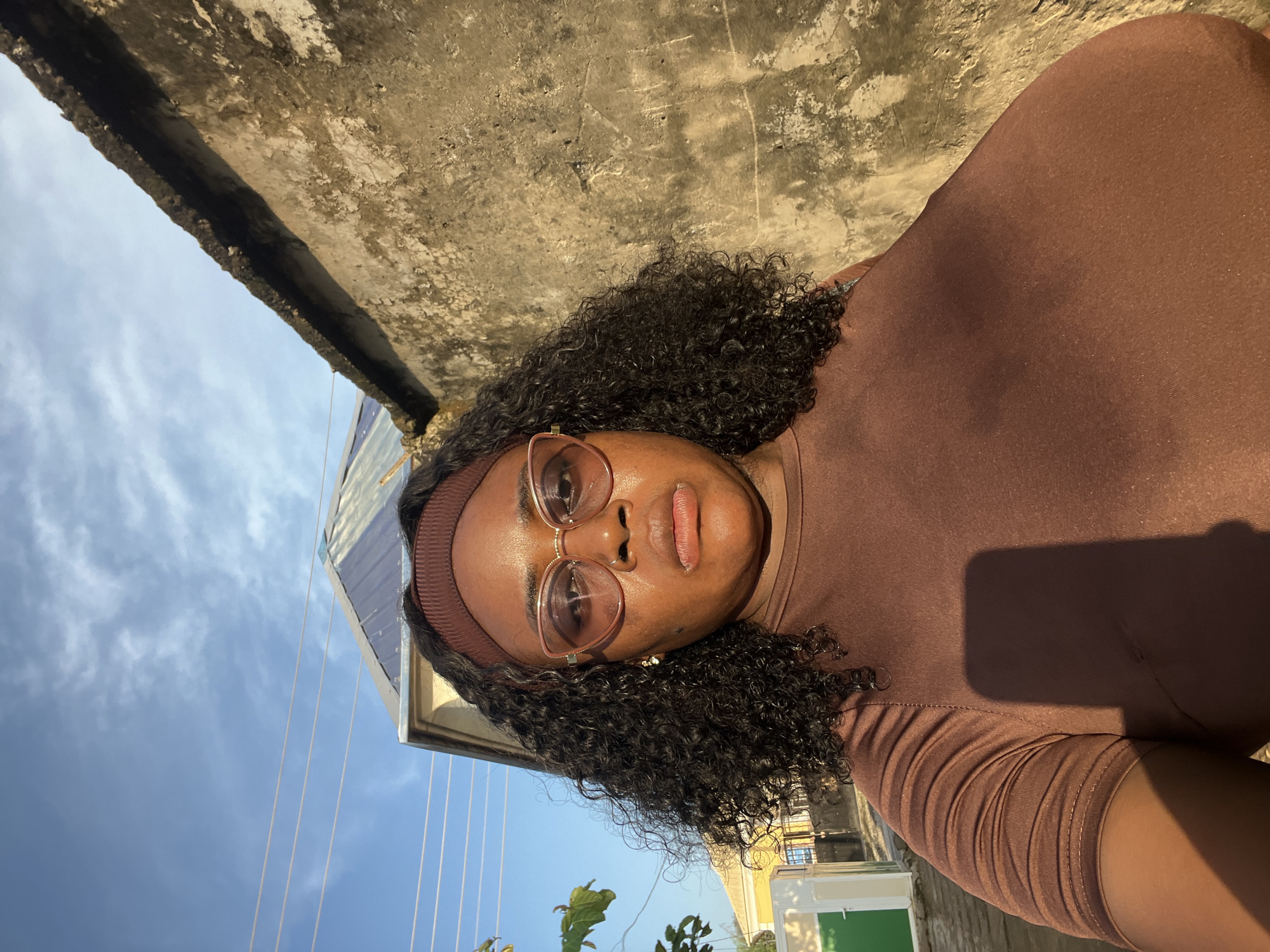 A self-portrait of a woman with dark, curly hair and glasses is taken outdoors. She is wearing a brown top and a brown headband. The background consists of a rough, weathered wall on the right, and a portion of a building with a metal roof on the left. The sky above is blue with some wispy clouds, and power lines are visible. The lighting suggests it is daytime, possibly late afternoon due to the angle of the shadows. The woman's facial expression is neutral. The scene appears to be in an urban or semi-urban environment, consistent with areas in Abuja, Nigeria.