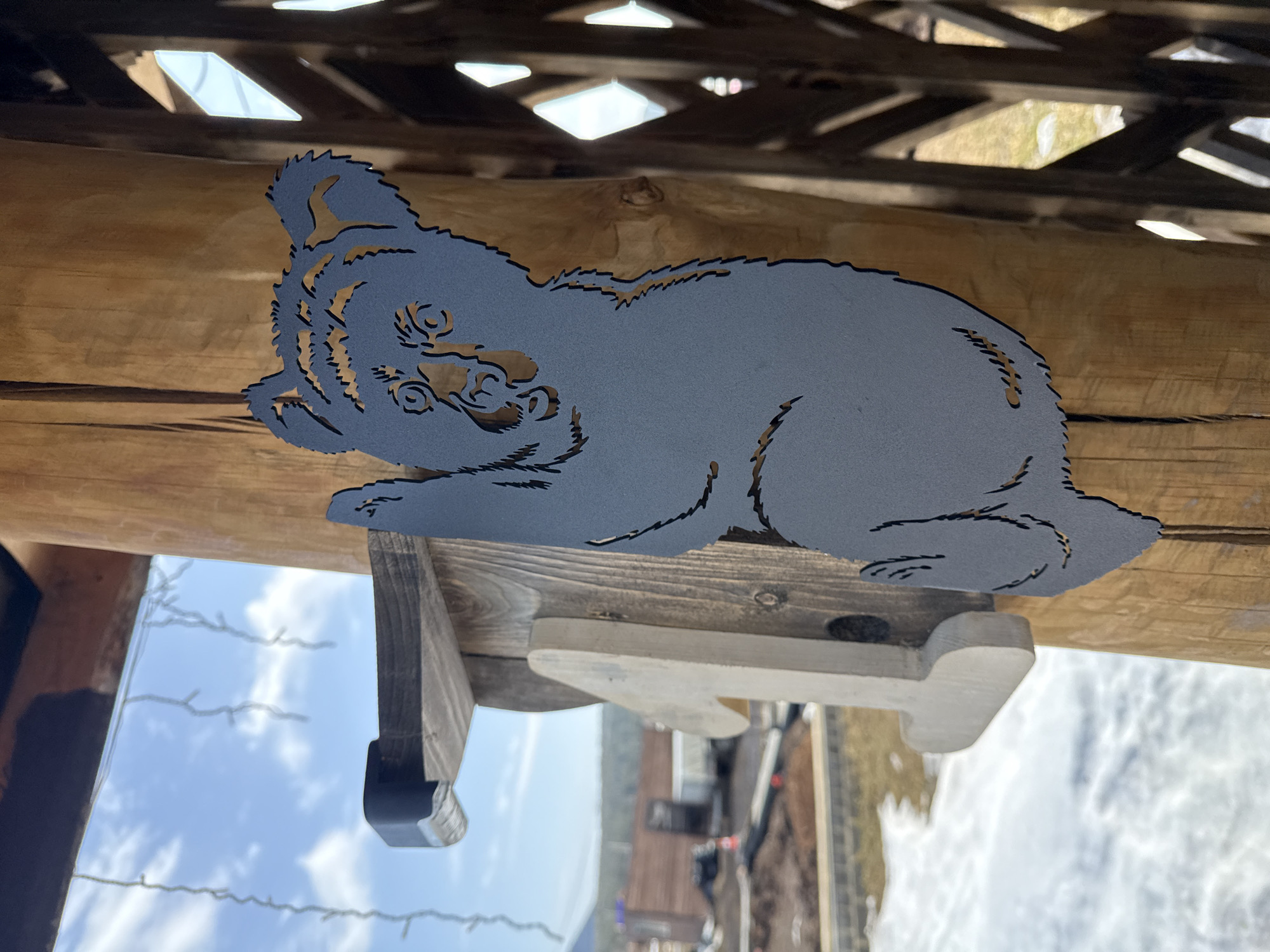 A gray, laser-cut metal silhouette of a bear cub, approximately 40-50 cm tall, is mounted on a vertical, light brown wooden post. The bear is depicted in a climbing posture, facing left. Below and to the left of the bear, a wooden cutout resembling the numeral "1" is attached to the same post, positioned below a black metal bracket. The post is part of a larger outdoor wooden structure, with a horizontal beam visible overhead and diagonal latticework on the right. The background reveals an outdoor landscape with patches of snow and exposed earth. In the mid-ground, a construction area is visible, featuring a reddish-brown building and a white vehicle or trailer. Further in the distance, obscured by a slight haze, are hills or mountains with visible tree lines. The sky is clear blue with scattered white clouds, and decorative string lights are suspended across the upper portion of the frame. The scene is located in Tyulyuk, Russia.