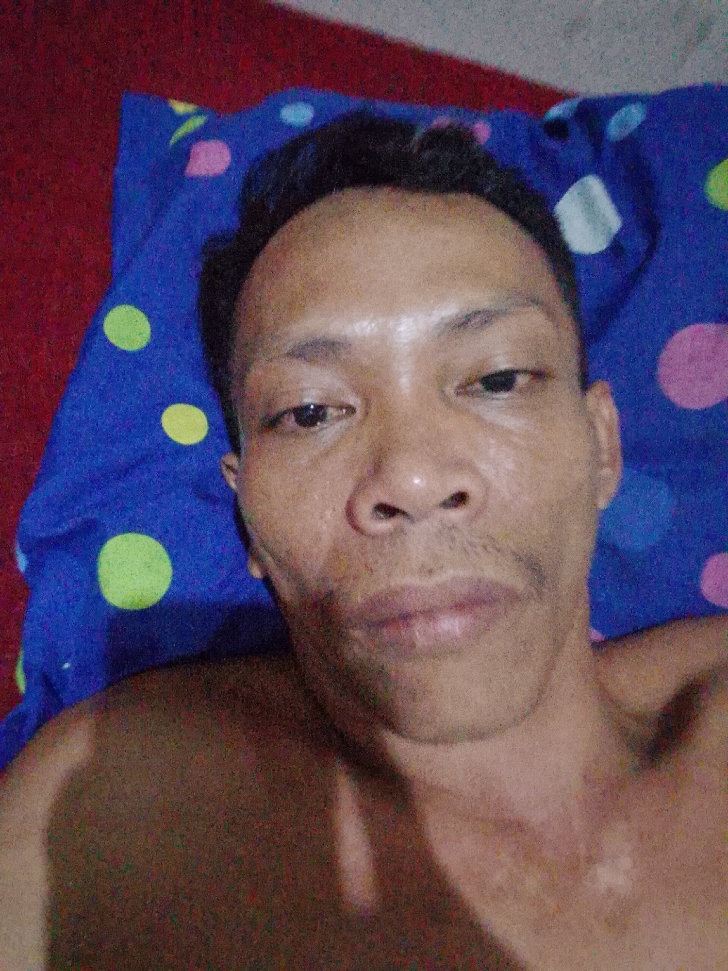 A medium close-up shot depicts a man with dark hair and medium brown skin tone, lying on a blue pillow patterned with multicolored circles (yellow, green, pink, and white). The background immediately behind the pillow appears red. The man's face is the primary focus, revealing details such as his eyes, nose, and mouth. His chest and shoulders are also visible. The image was likely taken indoors. There is no explicit information that specifies the exact location is Bukit Ketok, Indonesia.