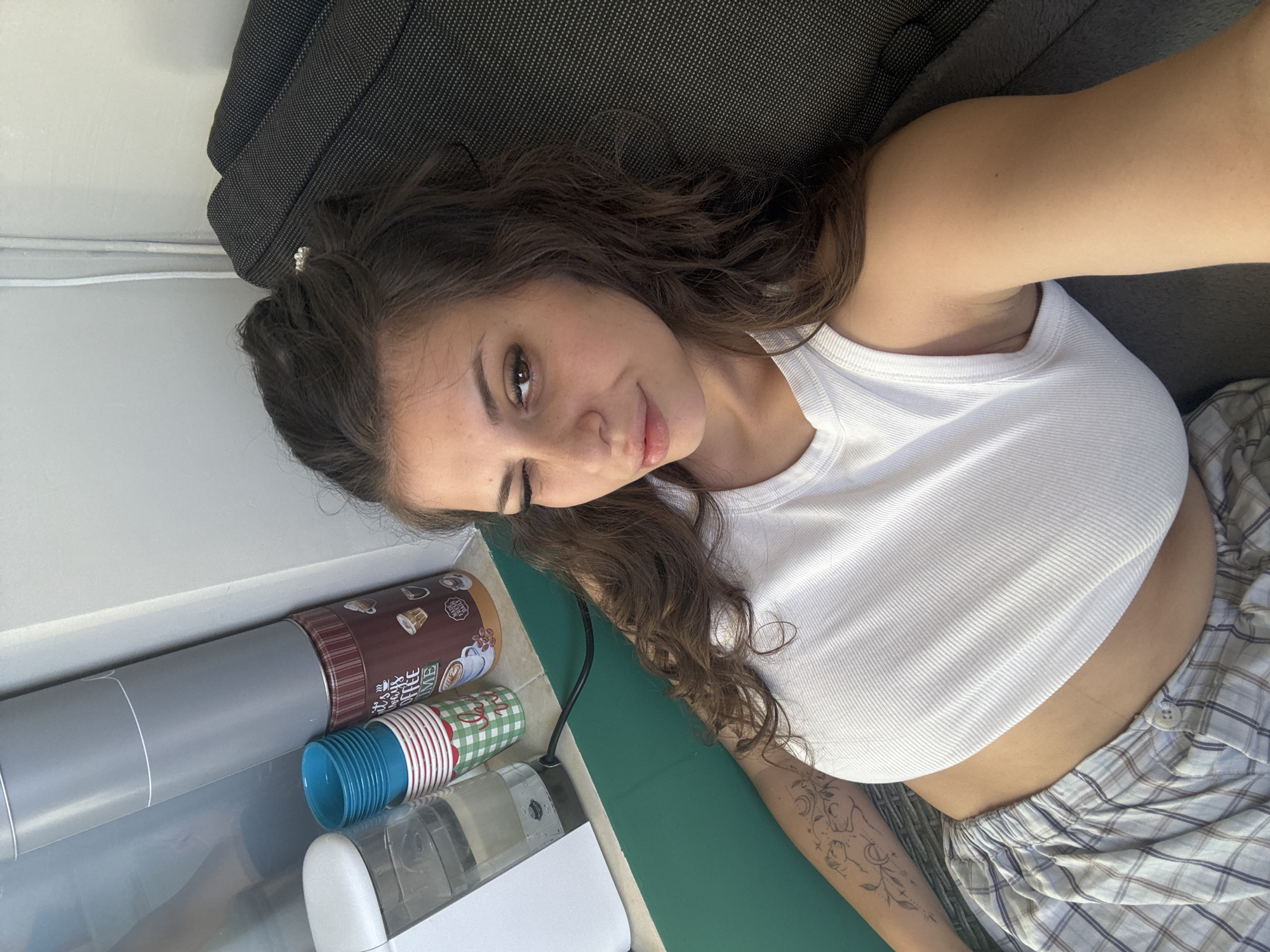 A woman is lying on a couch in what appears to be a living room. She is wearing a white tank top and plaid pajama pants. She is winking at the camera. There are some coffee cups and a canister of coffee on a table next to her. There is also a water dispenser and a green mat on the table. The room is lit by natural light coming from a window.