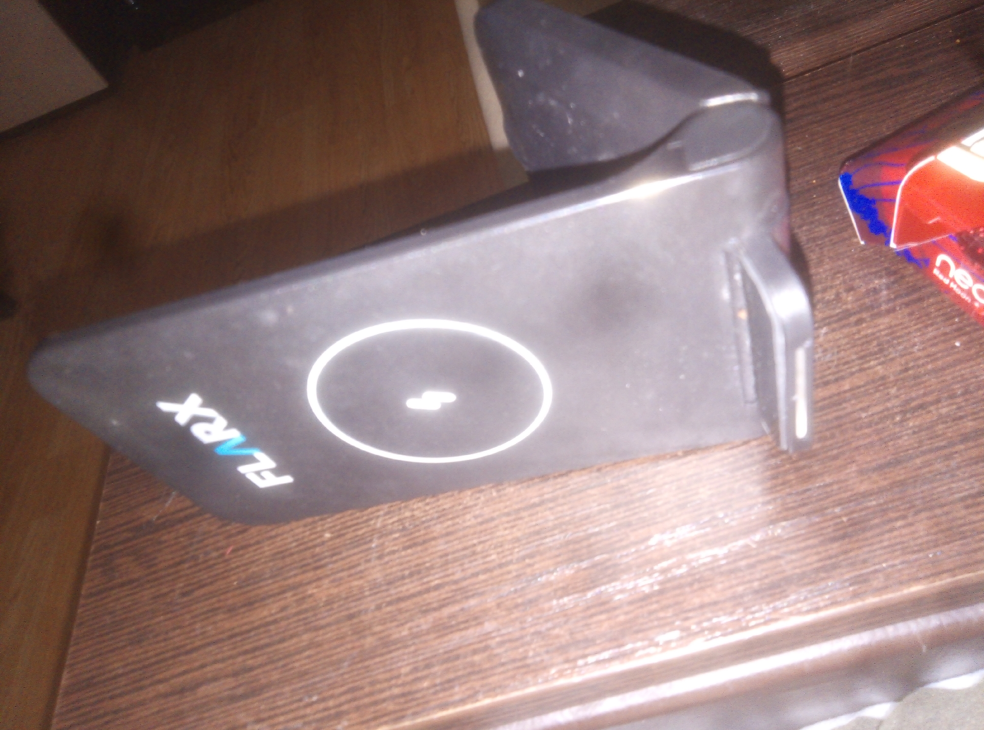 The image displays a black electronic device, likely a wireless charger or phone stand, positioned on a wooden surface. The device features a sleek, angular design with a flat base and an upward-sloping backrest. On its flat top surface, a prominent white circular outline is visible, enclosing a white lightning bolt symbol, which typically signifies a charging function. To the left of this symbol, the brand name "FLARX" is written in white letters, with the "AR" part subtly highlighted in a light blue color. A small, rectangular port or connector is visible on the right side of the device.
To the right of the black device, a red and blue box is partially visible. The word "neo" is clearly discernible in white text on this box, with smaller text underneath that reads "Red Hook." This box appears to be packaging for a consumer product, possibly related to heated tobacco. The wooden surface beneath the objects exhibits a distinct grain pattern, suggesting it is a table or desk in an indoor setting. The lighting suggests artificial illumination, casting some reflections on the device. No human activity or individuals are present in the scene, which appears static.