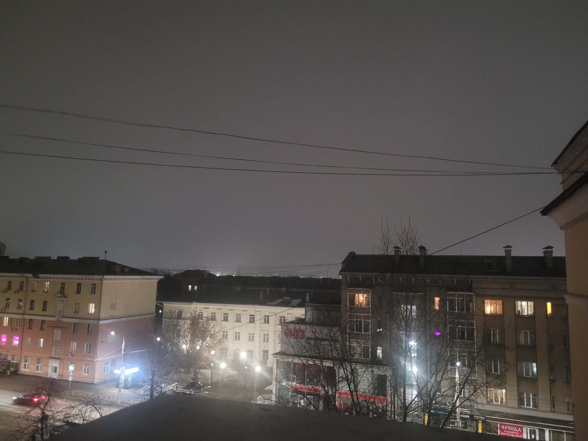 The image captures a nighttime cityscape in Izhevsk, Russia. It's a view from an elevated position, likely a high floor of a building, looking out over other buildings and streets. The sky is overcast and dark, with a few overhead wires stretching across the scene.
The buildings are primarily multi-story residential structures, typical of urban areas in Russia. Some have lights on in the windows, indicating activity inside. The architecture appears to be Soviet-era. The building to the left has a light-pink exterior, while the one to the right is primarily gray. There are bare trees in the foreground, suggesting it's a cold season, likely winter.
Streetlights illuminate the streets below, and there is a car driving on the road to the left, its headlights visible. The overall impression is of a quiet, dimly lit urban environment at night. A sign with the Russian word "APEHAA" is visible, which translates to "rent" in English.