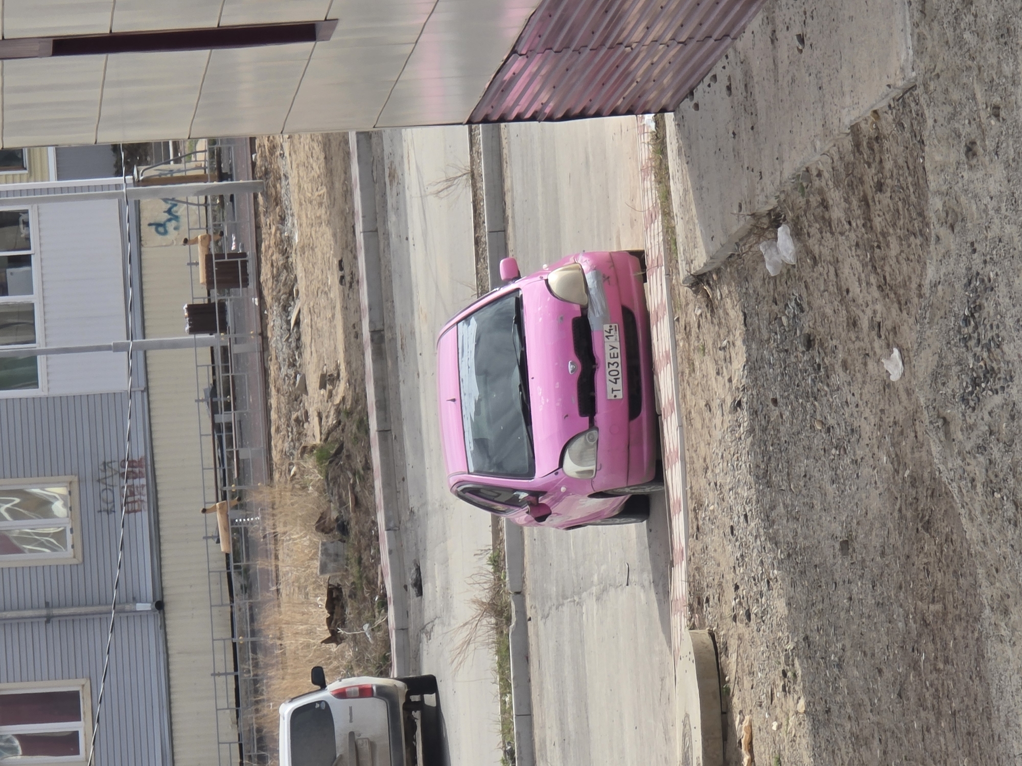 The image depicts a street scene in Lensk, Russia. A pink subcompact car is parked on the side of a road. The license plate of the car reads "T403EY 14." Adjacent to the road is a sidewalk and an unpaved area with gravel and sparse vegetation. In the background, there are buildings constructed with light-colored siding.