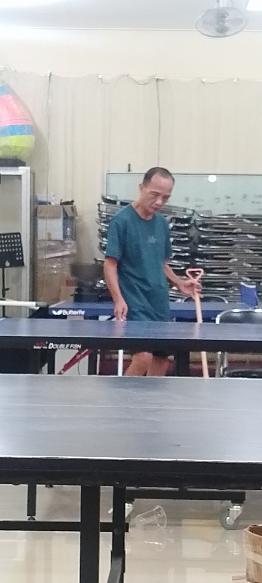 A middle-aged Asian man in a teal t-shirt and dark shorts stands behind a ping pong table. He is holding a wooden paddle with a pink handle. The man appears to be in an indoor facility, possibly a sports hall or gymnasium, given the presence of ping pong tables and a stacked display of what appear to be circular metal frames in the background. A ceiling fan is visible in the upper right corner. The overall lighting suggests an indoor environment, potentially during the day. The man's expression is neutral as he looks down at the paddle he is holding. The scene is clean and orderly, with no apparent signs of disorder or distress.