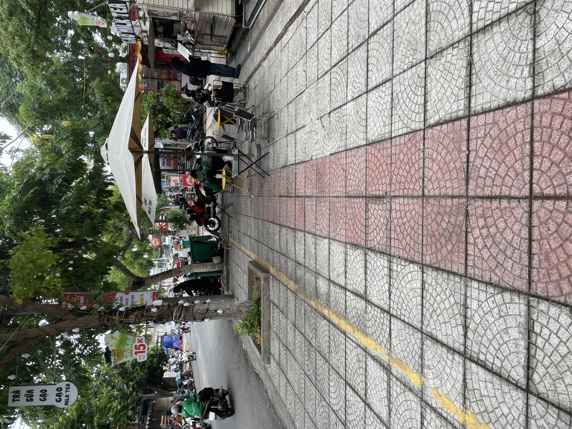 This image depicts a street scene in Da Nang, Vietnam, likely in a commercial area characterized by street-side businesses and pedestrian activity.
In the foreground, a textured sidewalk made of interlocking square tiles, with a central band of red tiles and a yellow painted line running along its edge, dominates the view. To the left, a road is visible with several motorcycles and scooters, some of which are occupied by individuals. A person in a green jacket is riding a black scooter.
Further into the scene, a sidewalk cafe area is set up with several tables and chairs situated under large, light-colored umbrellas. A small tree with decorative lights wrapped around its trunk stands in a planter on the edge of the sidewalk. Adjacent to this, a food stall, possibly a milk tea shop, is open for business, with customers and staff interacting. Signs indicating "GAO MILK TEA" are prominently displayed on the stall and on a banner hanging from a tree.
Several people are present, some seated at tables, others standing or walking. One person in a black hooded jacket is seen standing at the food stall, looking at a menu or order. Another person in a black t-shirt with "GAO MILK TEA" printed on it is seated at a table. A few other individuals are scattered around the cafe area and the food stall.
The environment appears to be a typical urban street setting with trees lining the sidewalk and buildings in the background. The weather seems overcast. The overall atmosphere suggests a lively commercial district with food and beverage services catering to passersby and those on motorbikes.