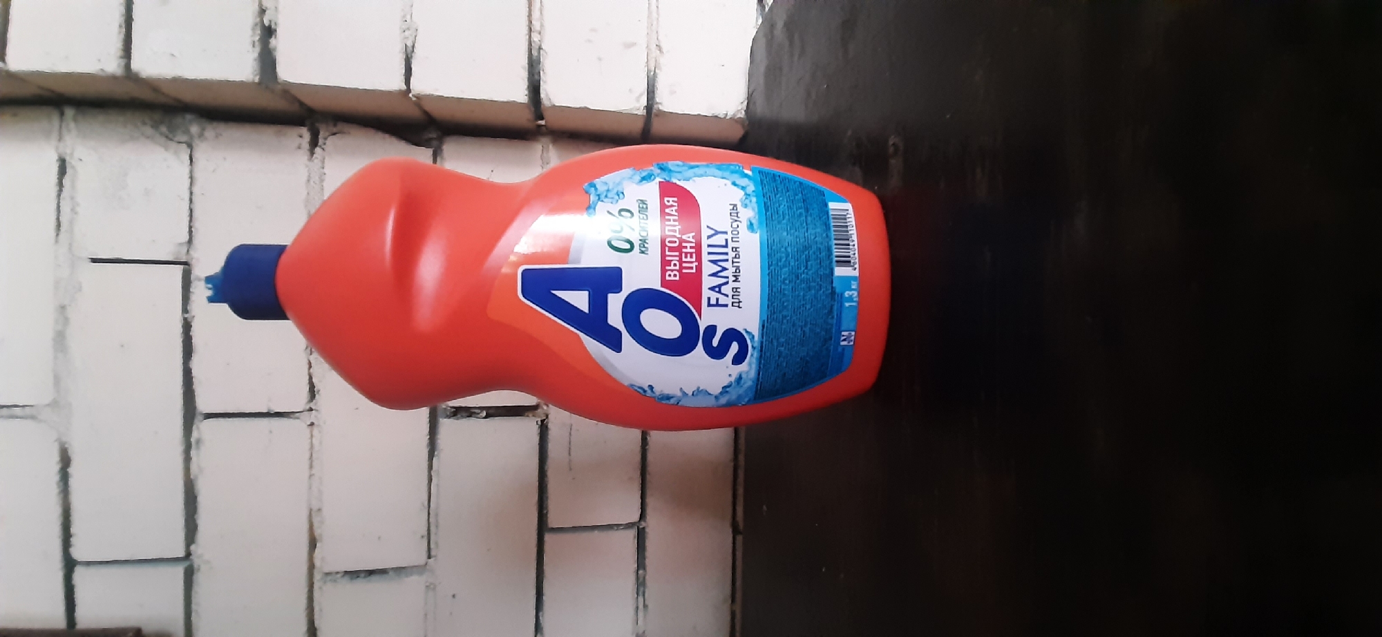 A close-up, eye-level shot shows an orange bottle of "AOS FAMILY" dish soap with a blue cap, standing on a dark, wooden surface. The bottle is positioned slightly off-center to the right. The label features large blue letters spelling "AOS," with smaller text below indicating "0% красителей" (0% dyes), "Выгодная цена" (Bargain price), and "Family для мытья посуды" (Family for dishwashing). Below this text, a barcode is visible, along with "1.3 л" (1.3 L). In the background, a white brick wall is visible, with a thin vertical dark line to the right of the bottle. The scene is indoors, and the lighting appears to be natural. The city of Yessentukskaya, Russia, is mentioned in the prompt, but the image itself does not provide any visual cues to confirm or deny this location.